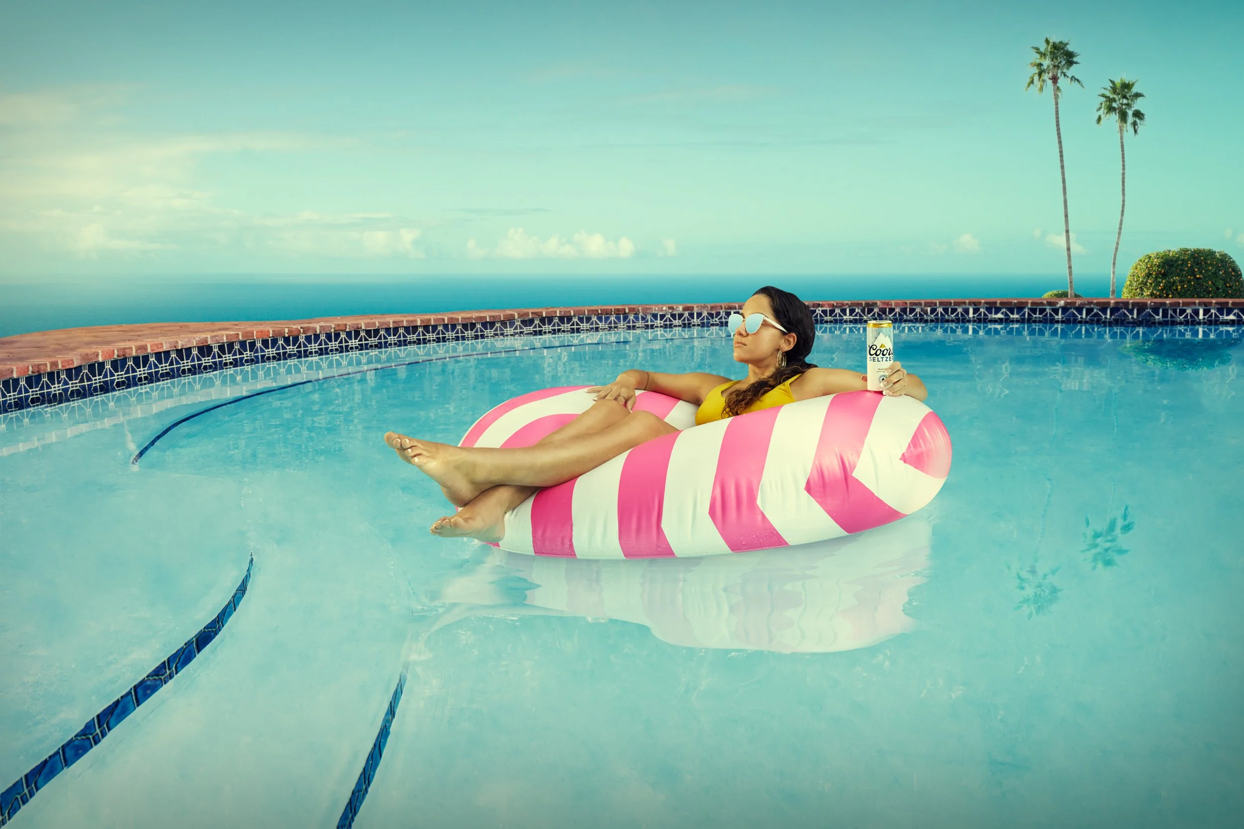 Woman lounging on a pink and white float in a swimming pool, holding a can of Coors Seltzer, wearing sunglasses and a yellow swimsuit, with palm trees and the ocean in the background.