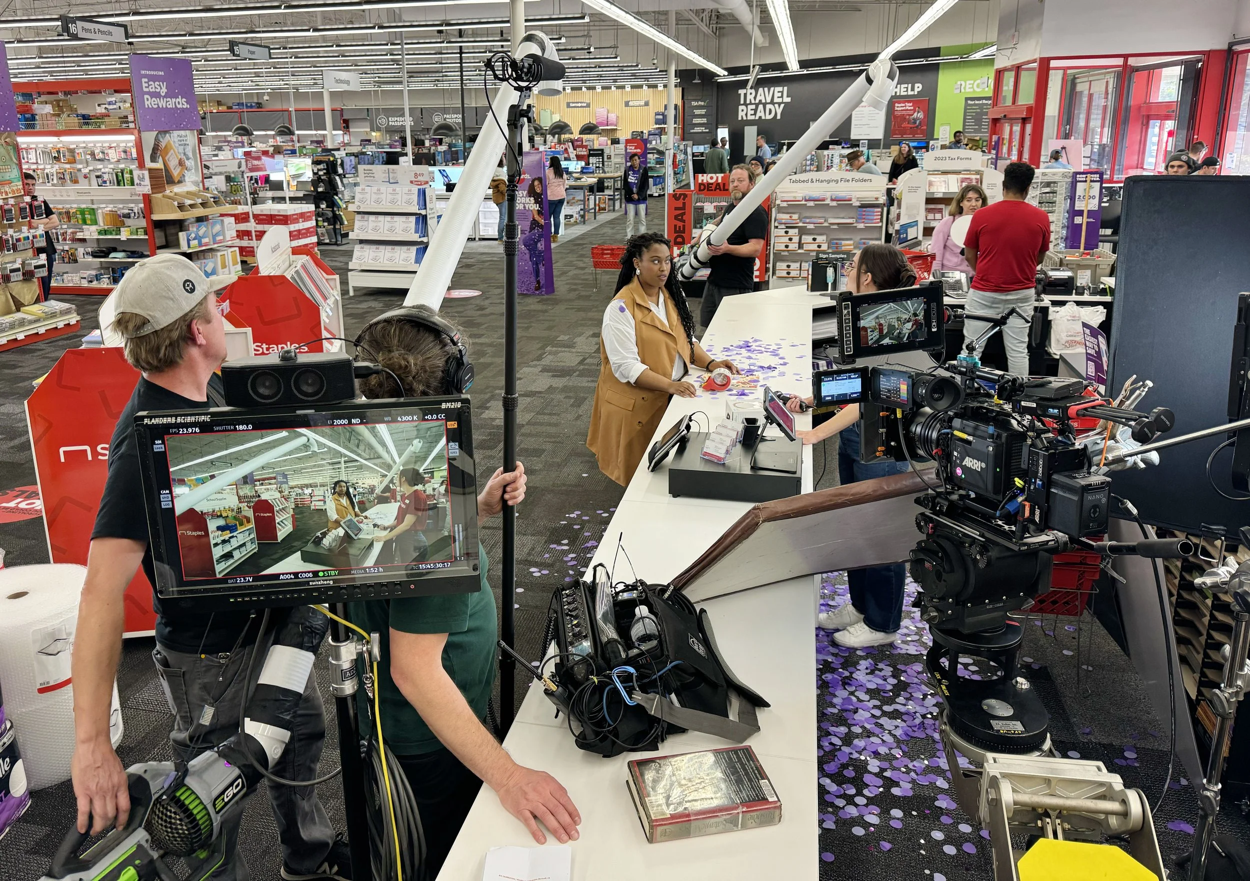 A scene inside a retail store, likely a Staples, with a filming crew filming a woman at a checkout counter. The store has various aisles with office supplies and promotional signs visible in the background. The floor is covered with purple confetti.
