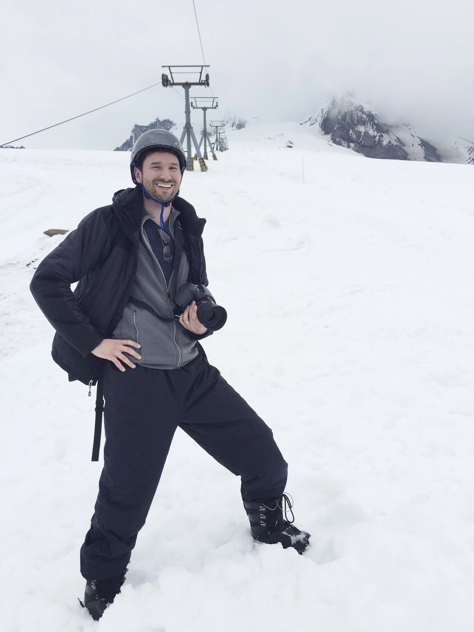 A man standing in snow on a mountain with a camera around his neck, smiling, wearing a helmet, black jacket, gray hoodie, black pants, and snow boots, with ski lift towers and mountain peaks in the background.