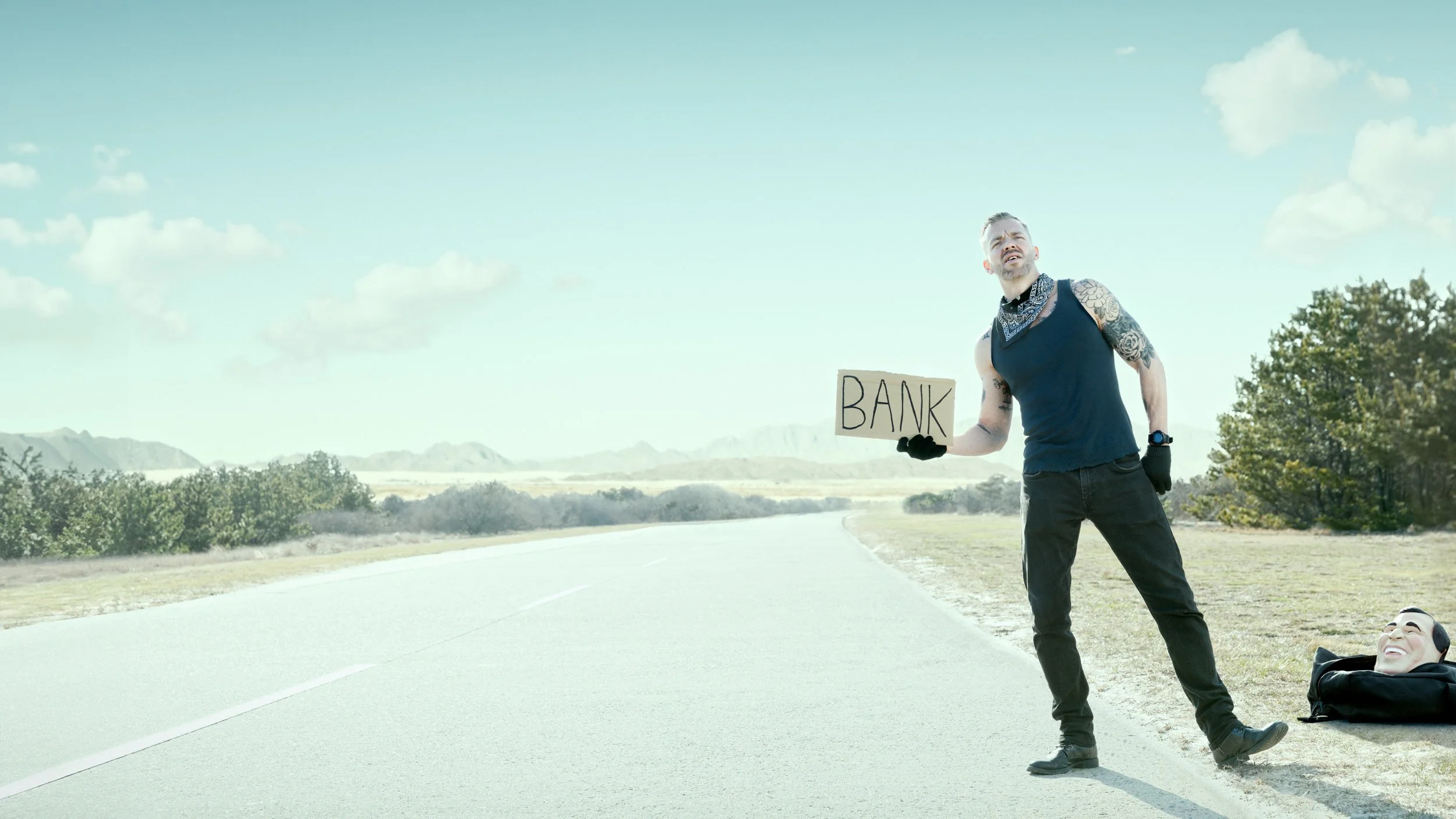 A man with tattoos wearing black gloves and a blue bandana around his neck holding a cardboard sign that says 'BANK' stands by the side of an empty road in a rural area with trees and mountains in the distance. A mask with a smiling face is lying on 