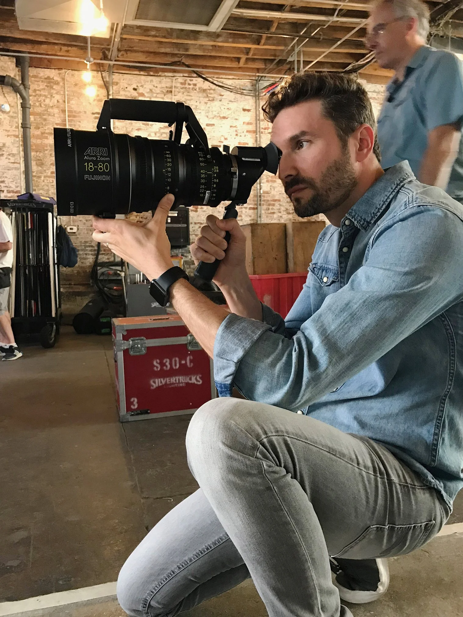 A man in a denim shirt crouches and looks through a large professional film camera with a long lens in an industrial setting.