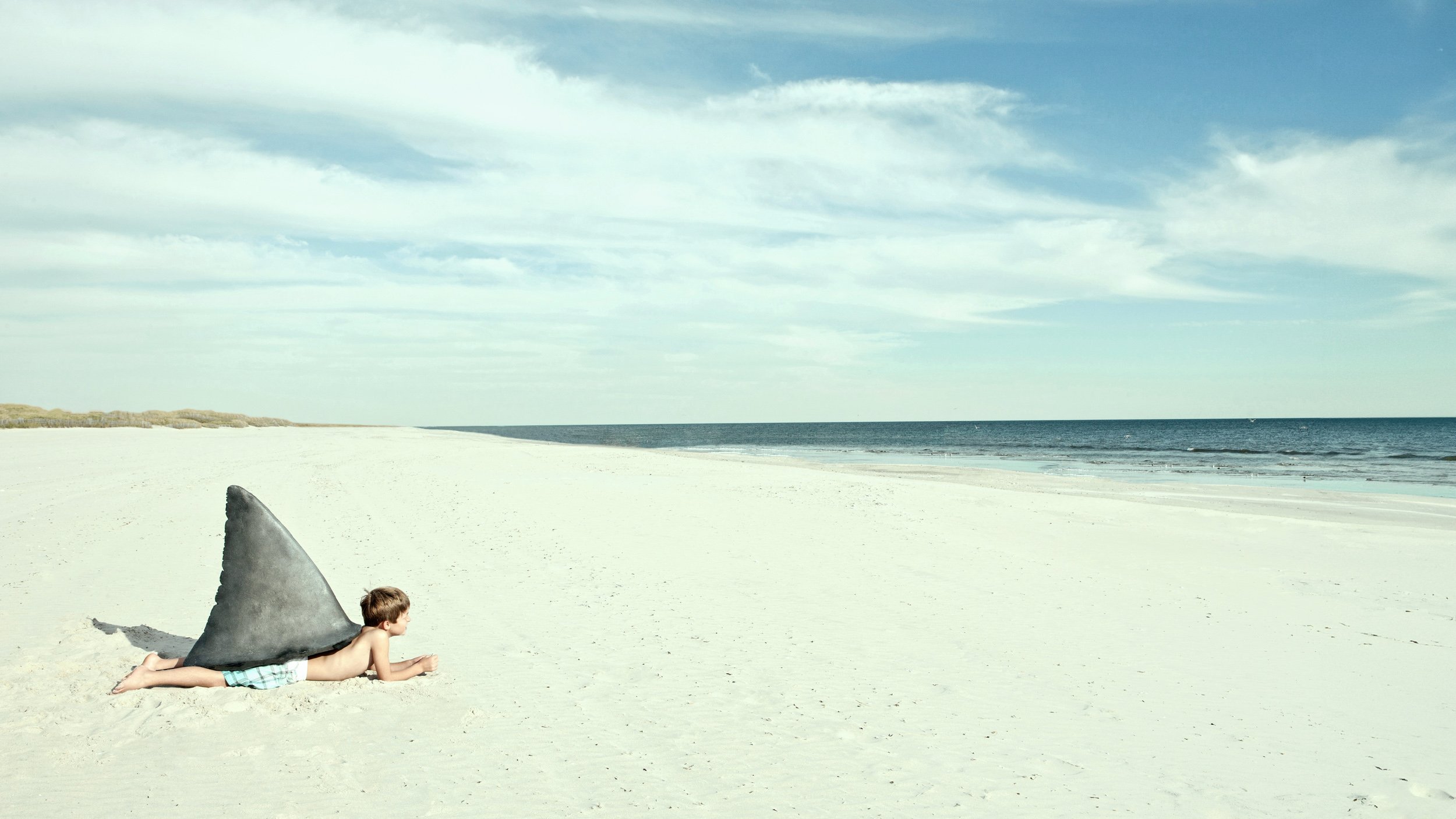 A young boy lying on a sandy beach, wearing swim trunks and a shark fin costume on his back. The beach is empty with the ocean and sky in the background.