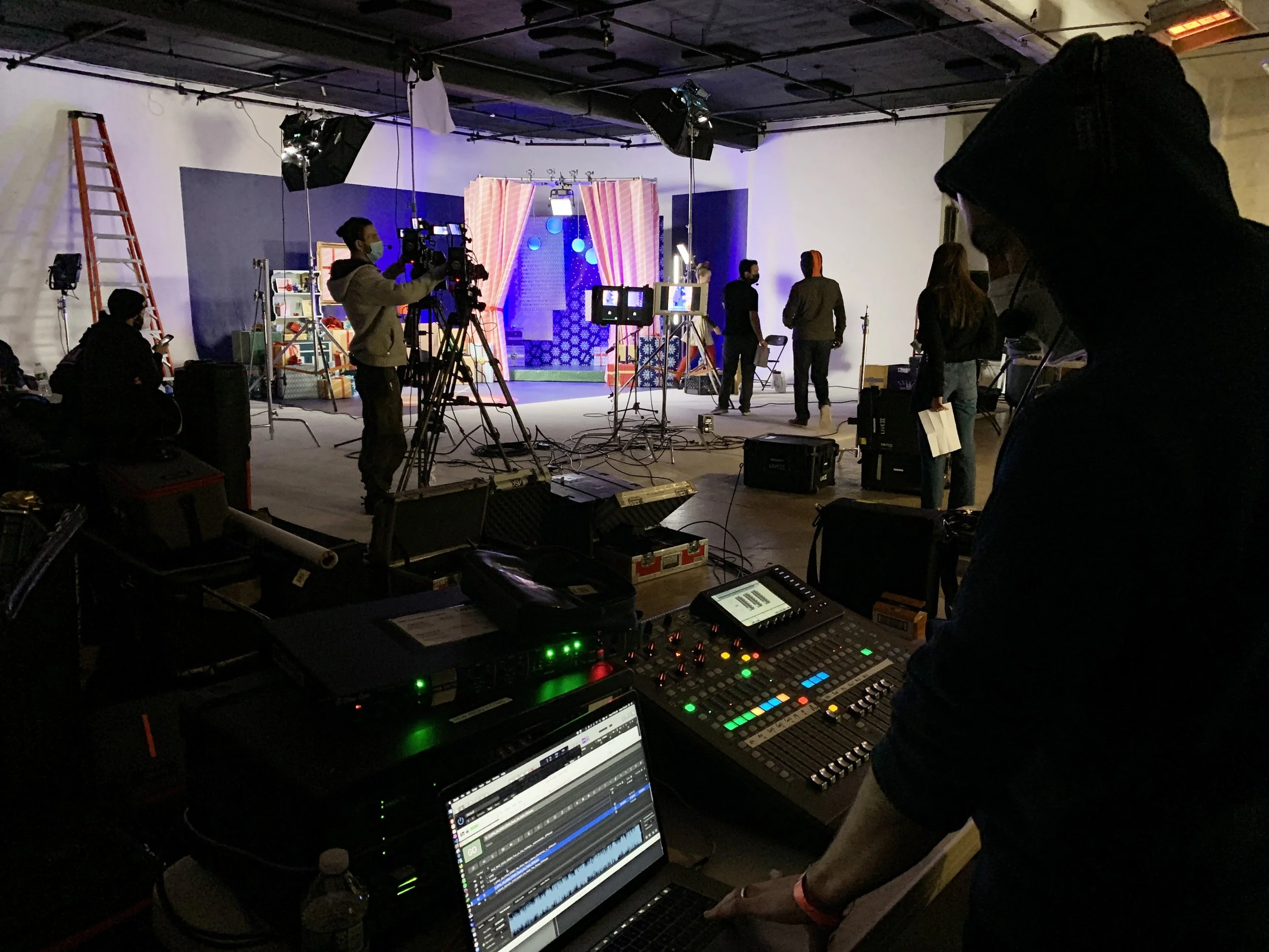 Behind the scenes of a video or film production in a studio, with camera operators, crew members, and a set with curtains and colorful background elements.