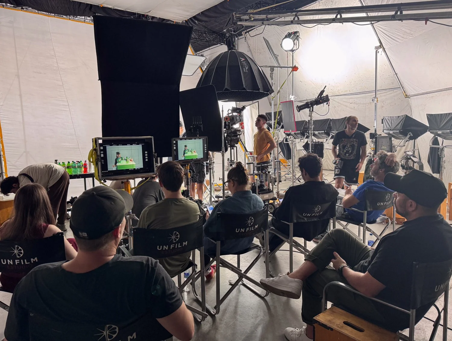Film crew on set filming a scene with lighting and camera equipment, and people seated in chairs watching the process.