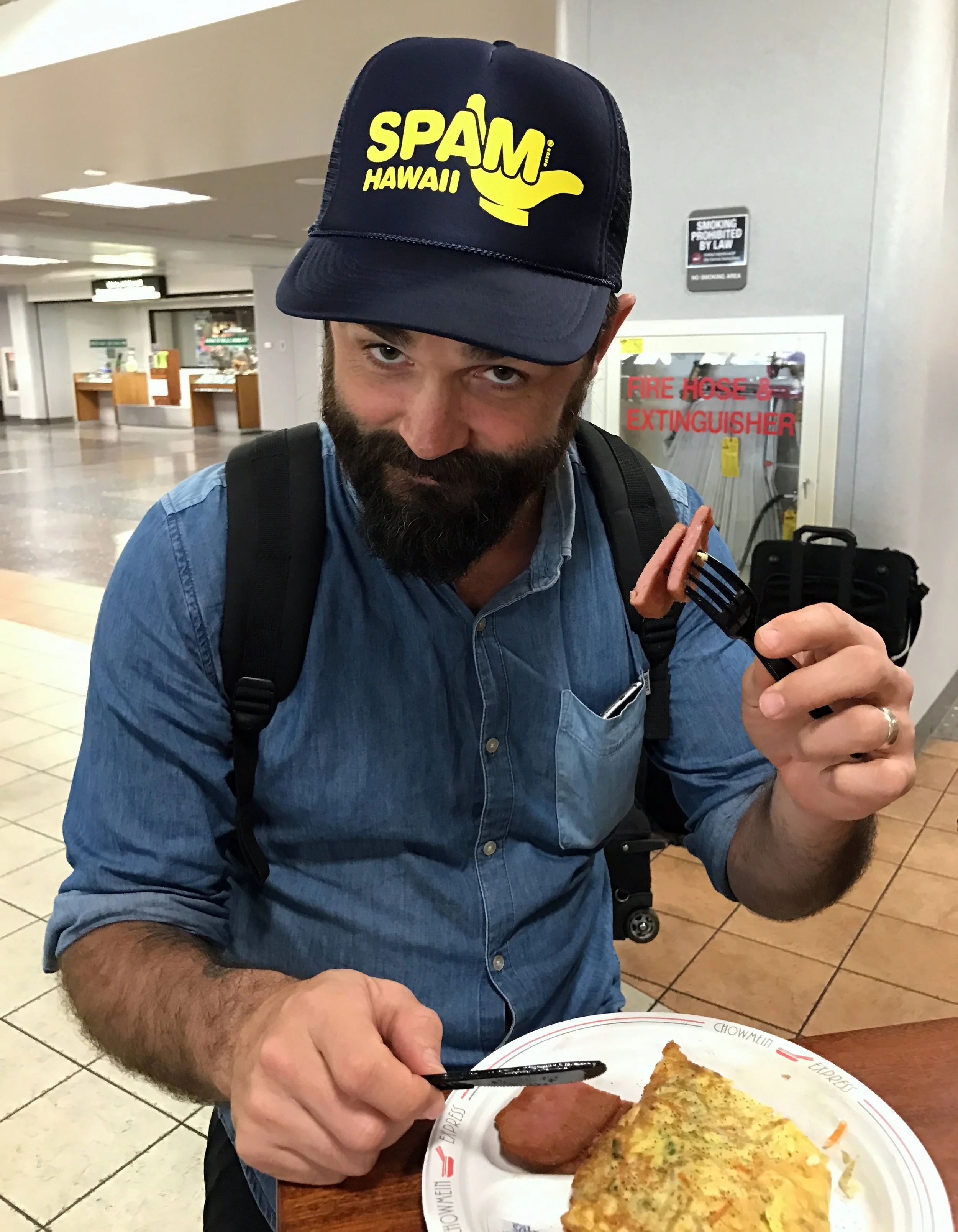 A man with a beard and blue shirt wearing a navy baseball cap with yellow logo and text reading 'Spam Hawaii' holds a fork with a sausage and a plate of food including an omelette and sausage at an airport dining area.
