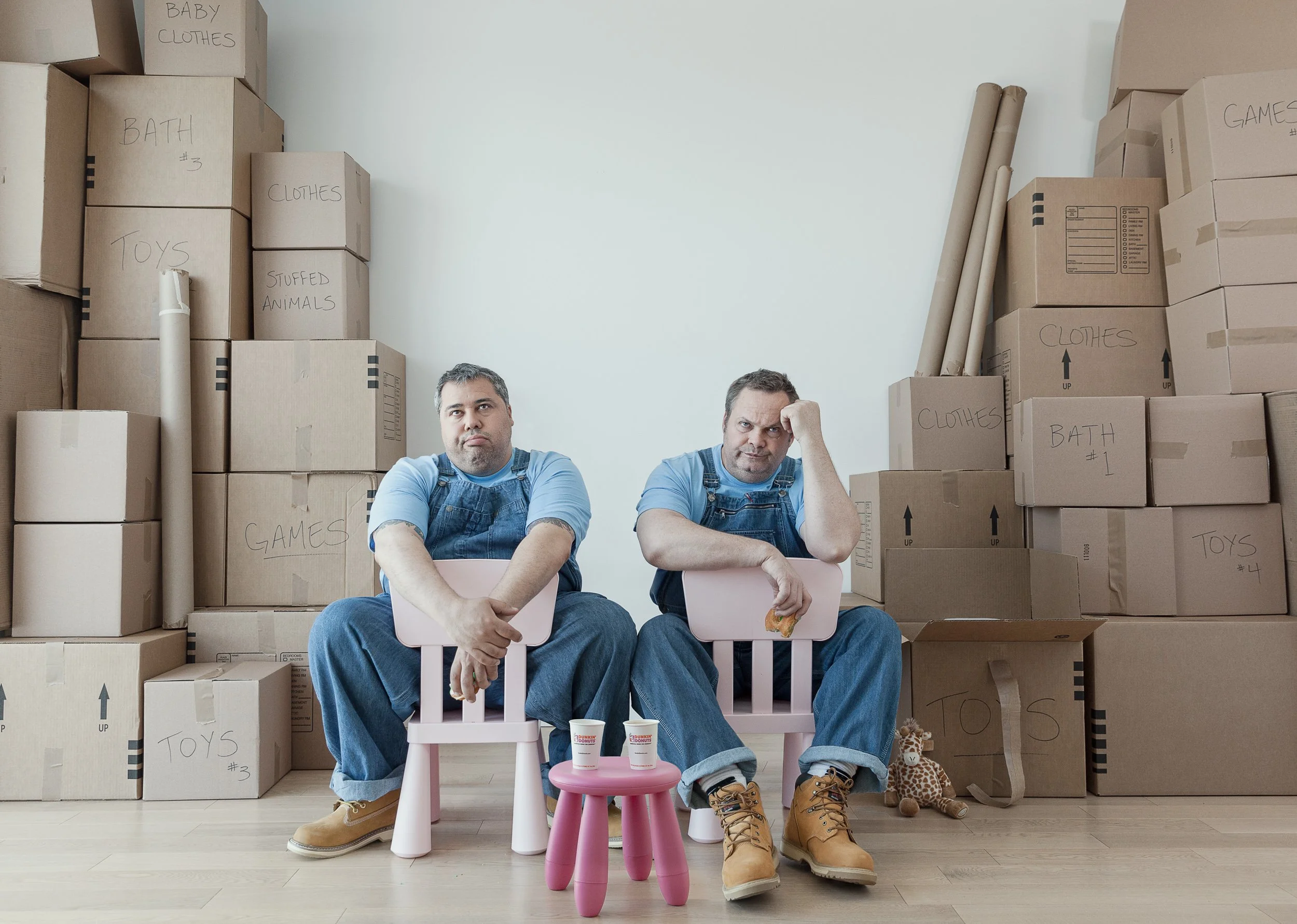 Two men sitting on pink chairs in front of a wall of cardboard boxes labeled with room names and contents, wearing blue shirts and jeans, with a small pink table holding two cups between them, and toys including a stuffed giraffe on the floor.