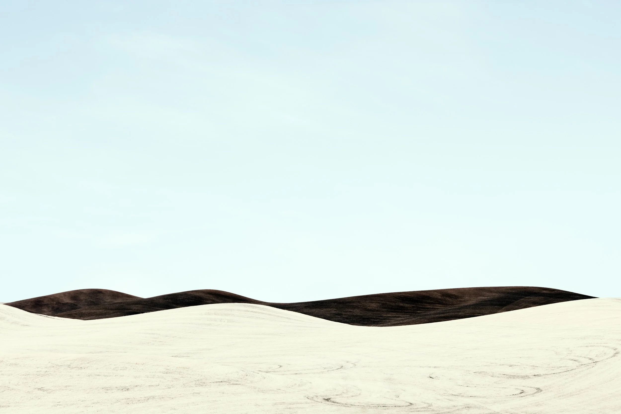 A desert landscape with light-colored sand dunes in the foreground and dark, rolling hills in the background under a pale sky.