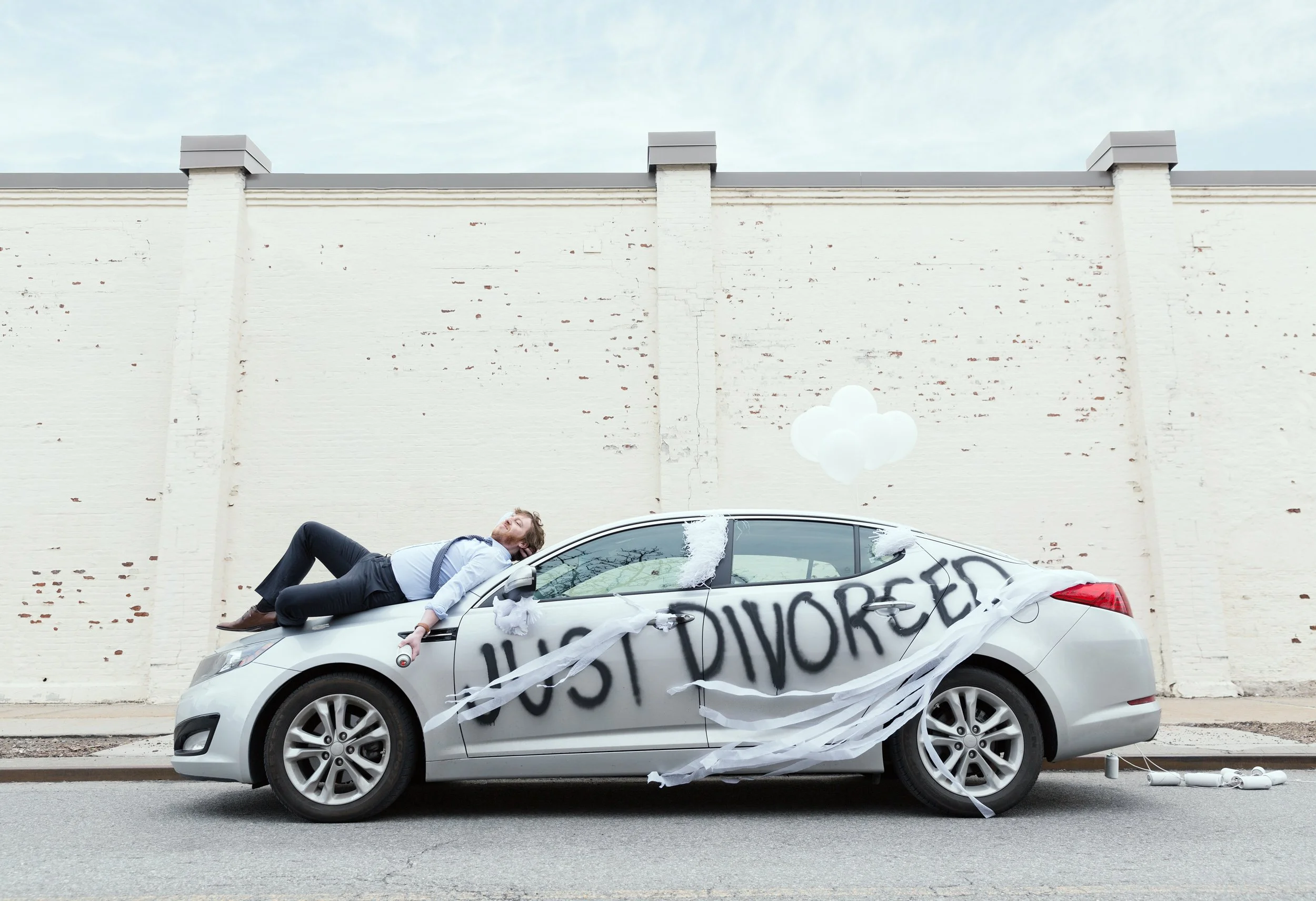 A silver car with a large banner reading 'LOST DIVORCED' and white decorations, with a man lying on the hood, appearing to be asleep or unconscious, in front of a white brick wall and cloudy sky.