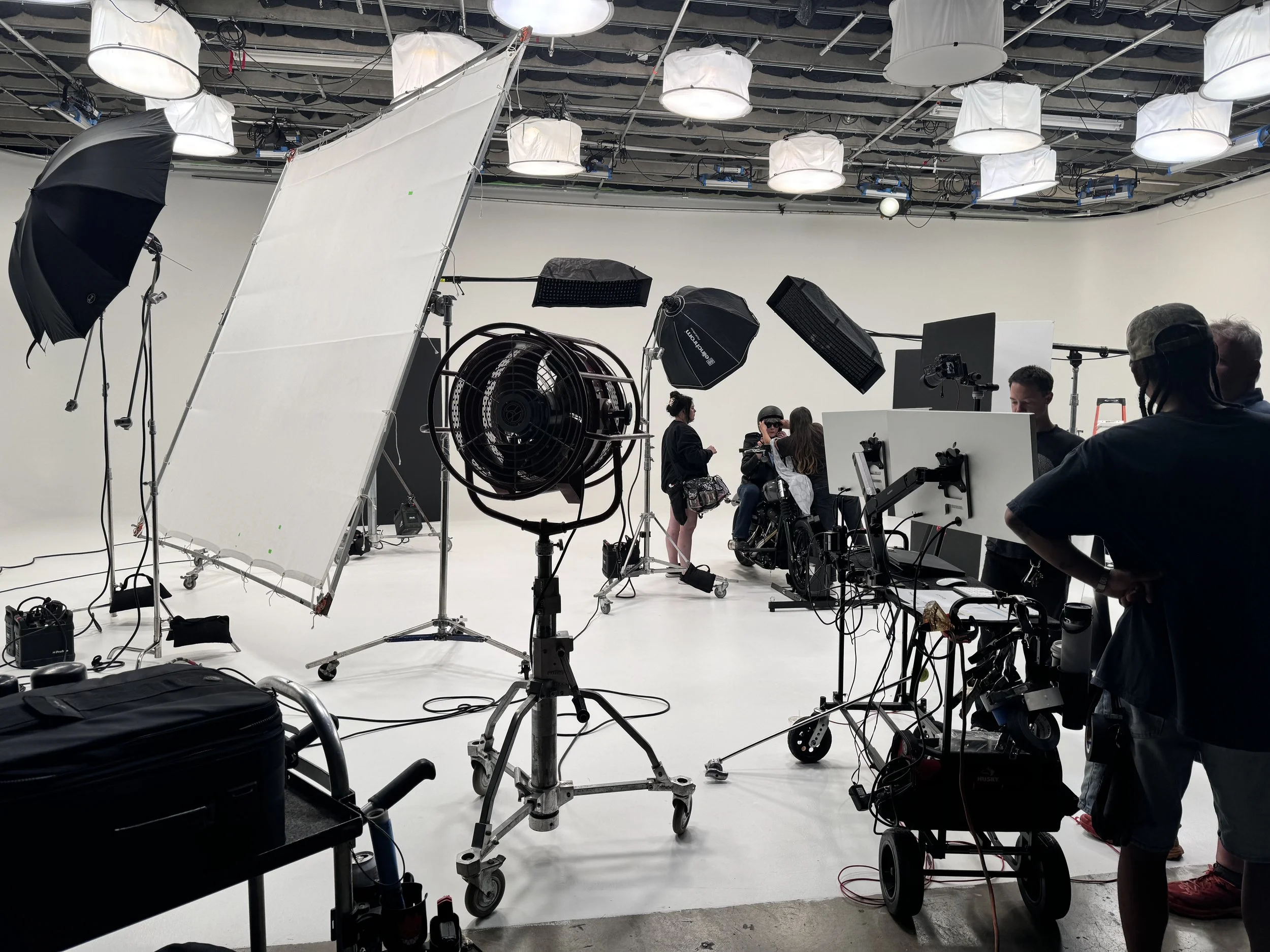 Film crew preparing a scene in a white studio with lighting equipment, a large fan, and a white backdrop, with three people sitting on a camera dolly.