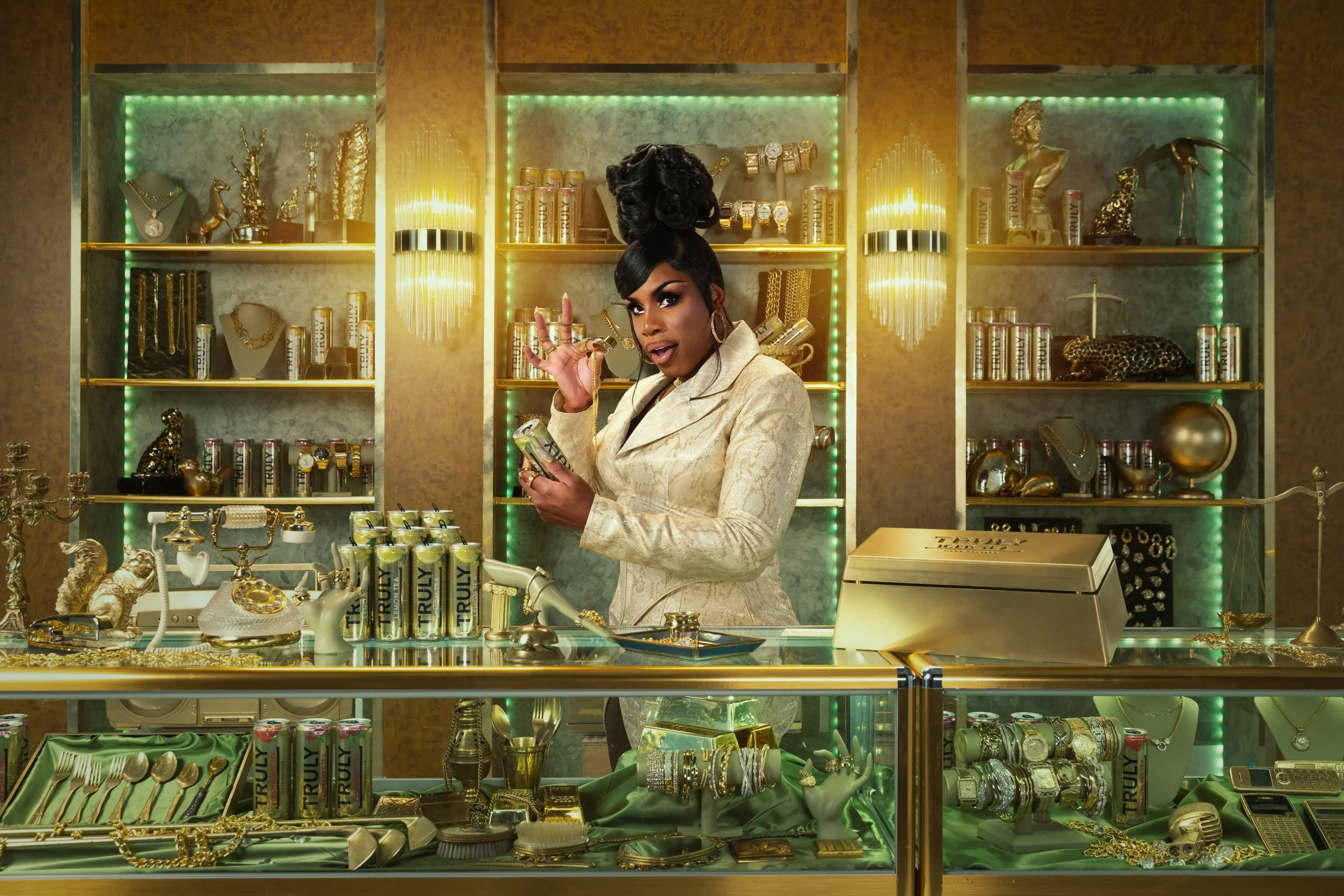 A woman with dark hair and makeup in a cream-colored blazer inside a luxurious jewelry and accessories store with gold accents, shelves of jewelry, watches, and decorative items, and a glass display case with jewelry and gold cans labeled 'Truly'.