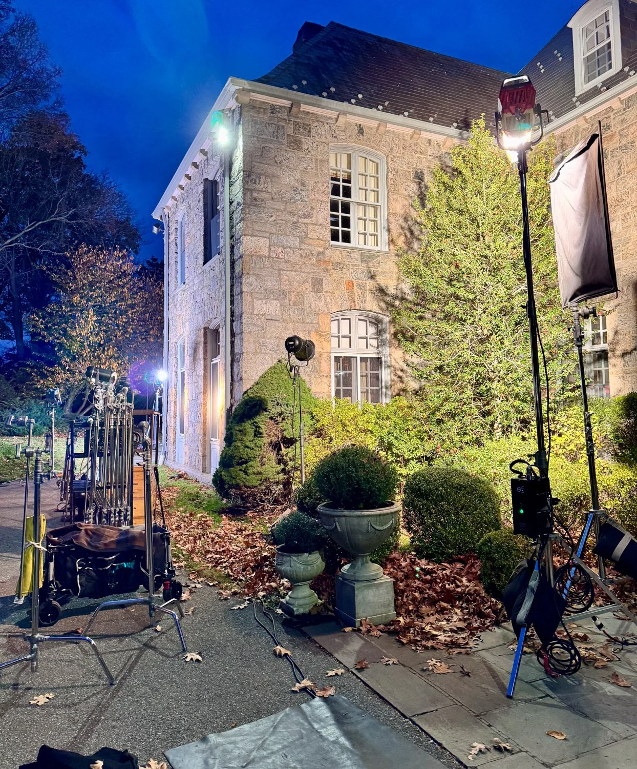 Behind the scenes of a film shoot outside a stone house at night, with lighting equipment, cameras, and crew setup.
