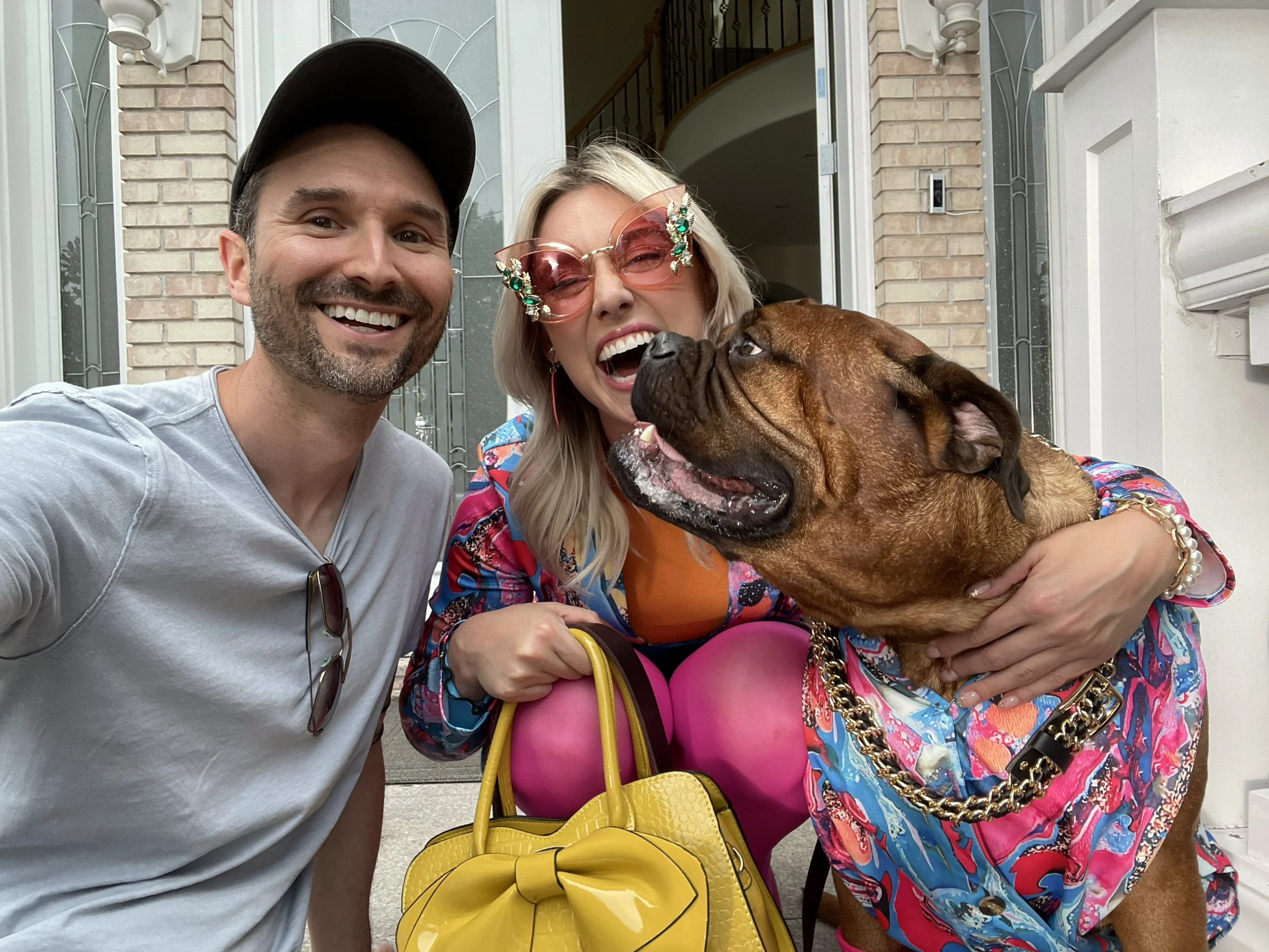 Three people and a dog taking a selfie in front of a house door. The man on the left has short dark hair, a beard, and is wearing a light gray shirt and a black cap. The woman in the middle has blonde hair, pink sunglasses decorated with green jewels