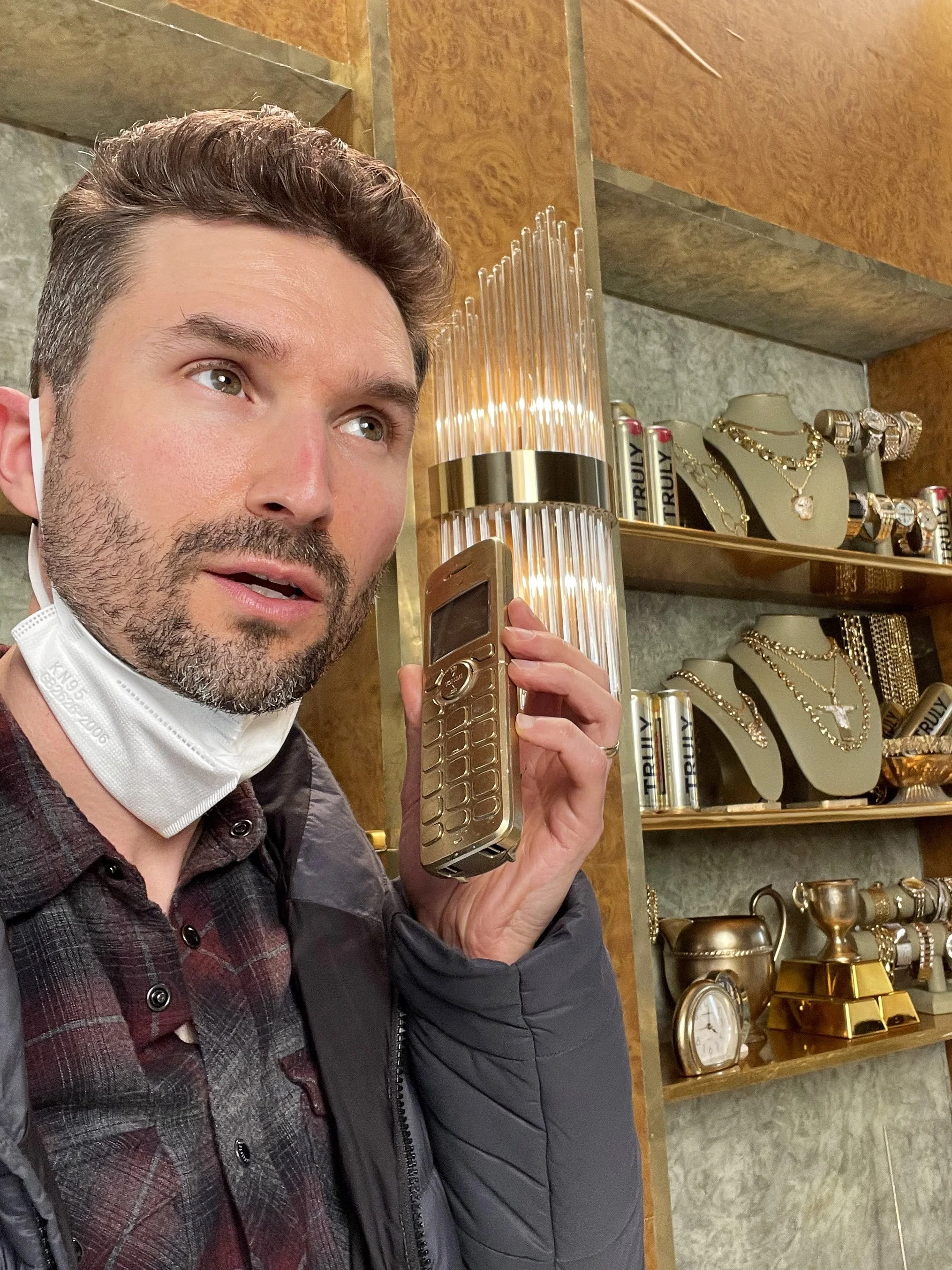 A man with a beard and checkered shirt holding an old mobile phone to his ear with a decorated jewelry display in the background.