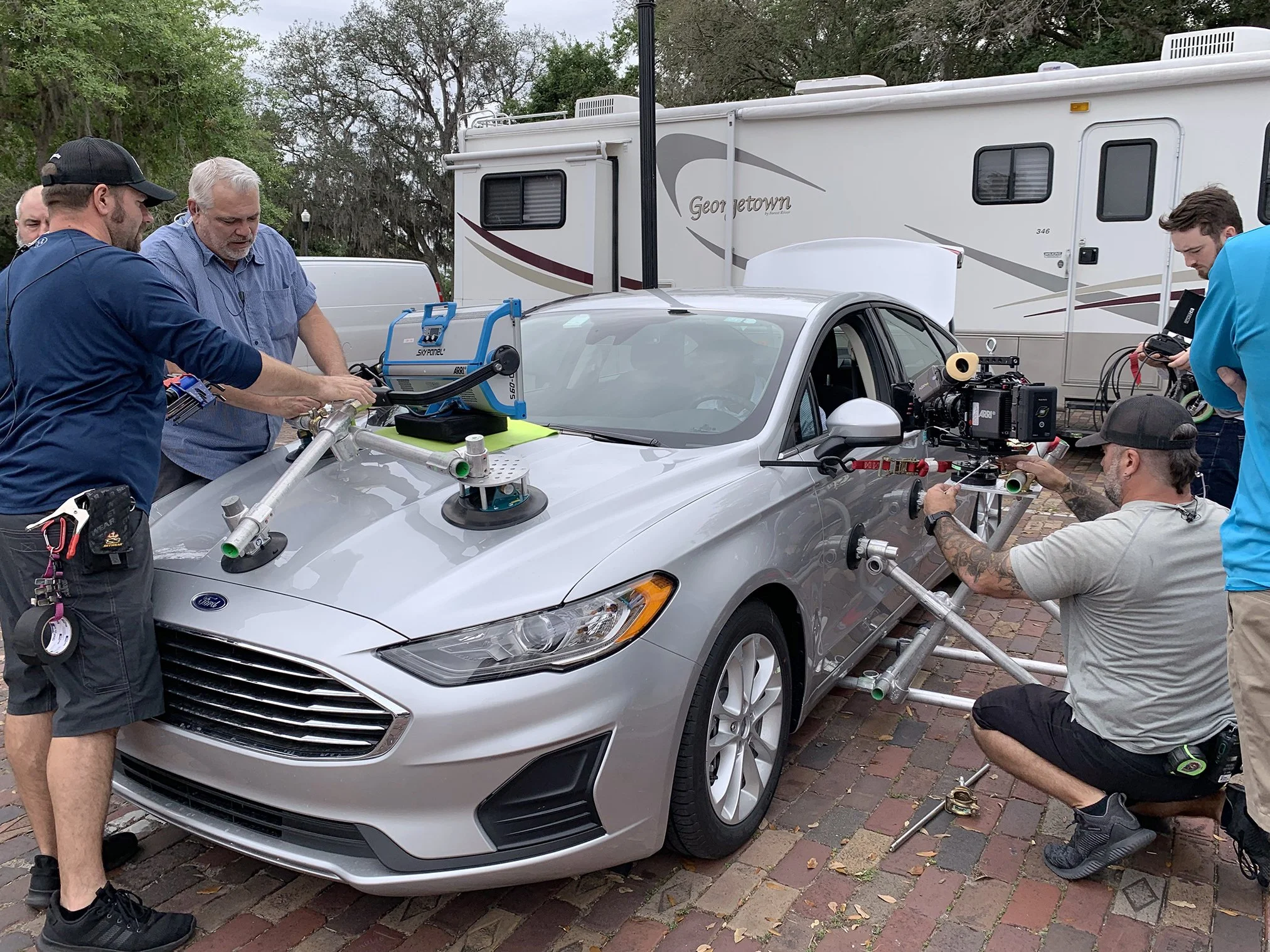 Film crew filming a silver Ford sedan car outdoors with crew members working on camera equipment and vehicle.