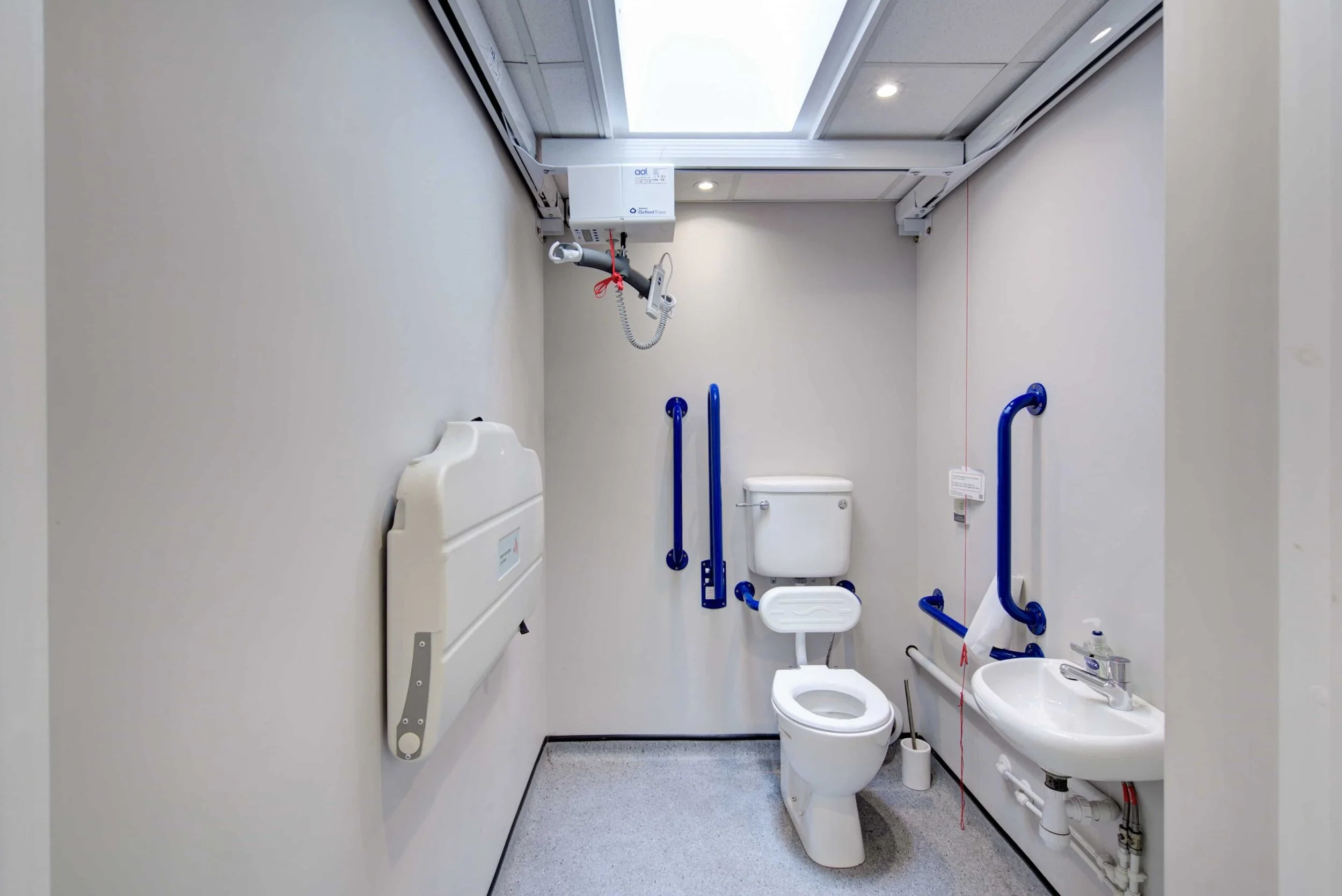 Accessible bathroom with a toilet, sink, blue grab bars, and medical equipment mounted on the ceiling for assistance.