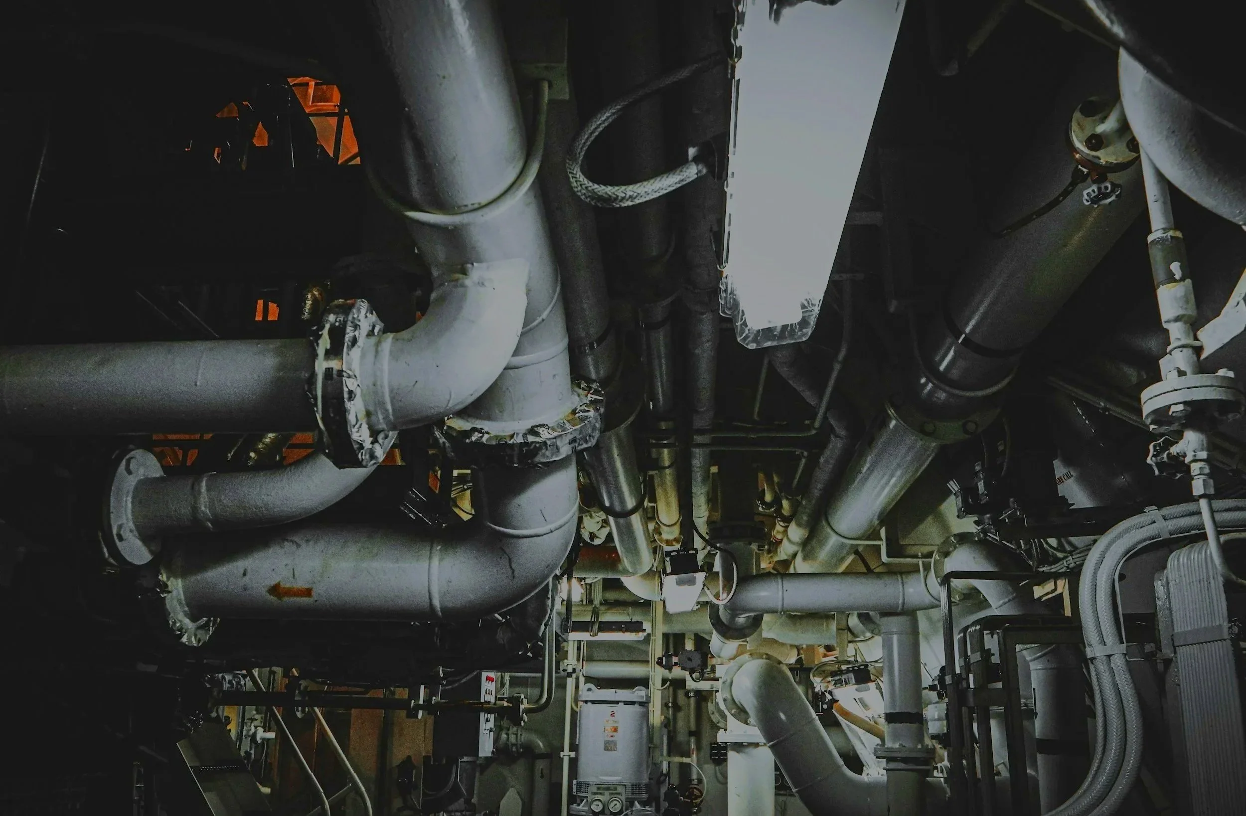 Complex network of gray and metallic pipes and valves within an industrial or mechanical room.