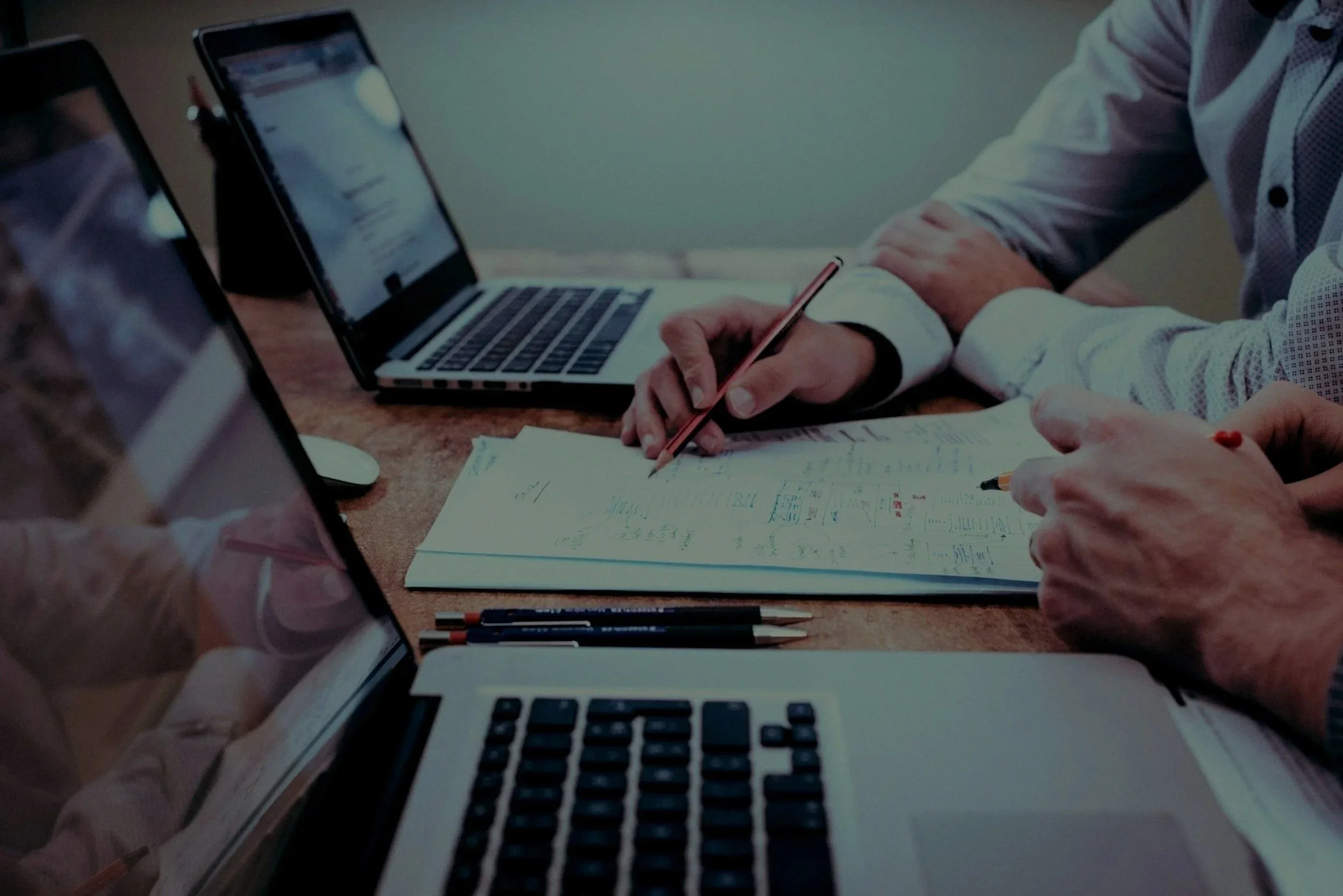 Two people working at a desk with laptops, papers, and pens, indicating a collaborative work or meeting.