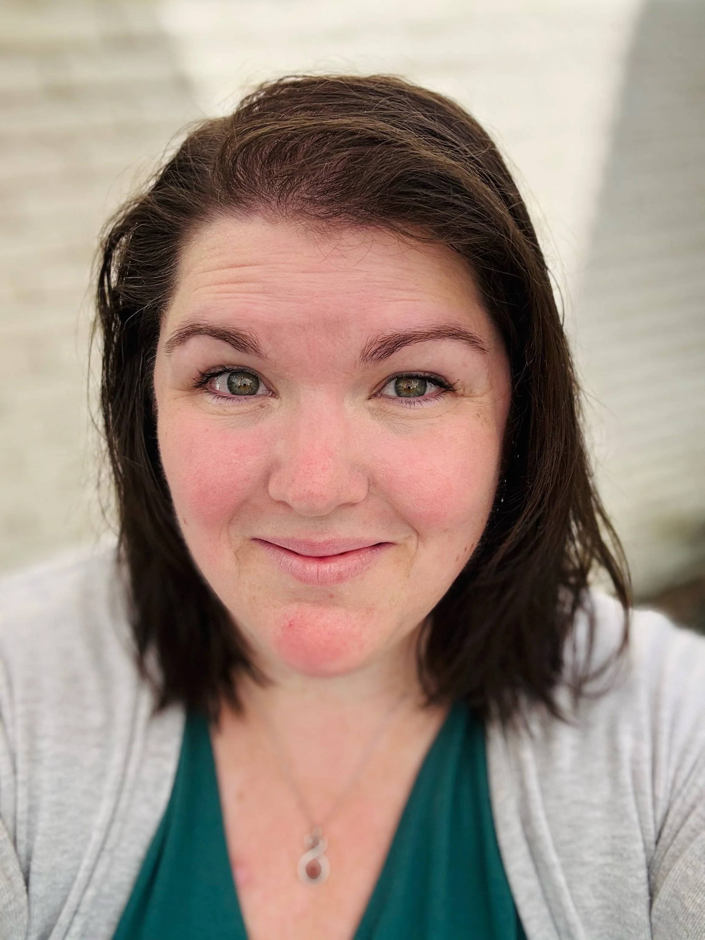 A woman with shoulder-length dark brown hair, light skin, and green eyes is smiling at the camera. She is wearing a green top, a light gray cardigan, and a silver necklace with a teardrop-shaped pendant. The background is blurred with what appears to be a light-colored wall.