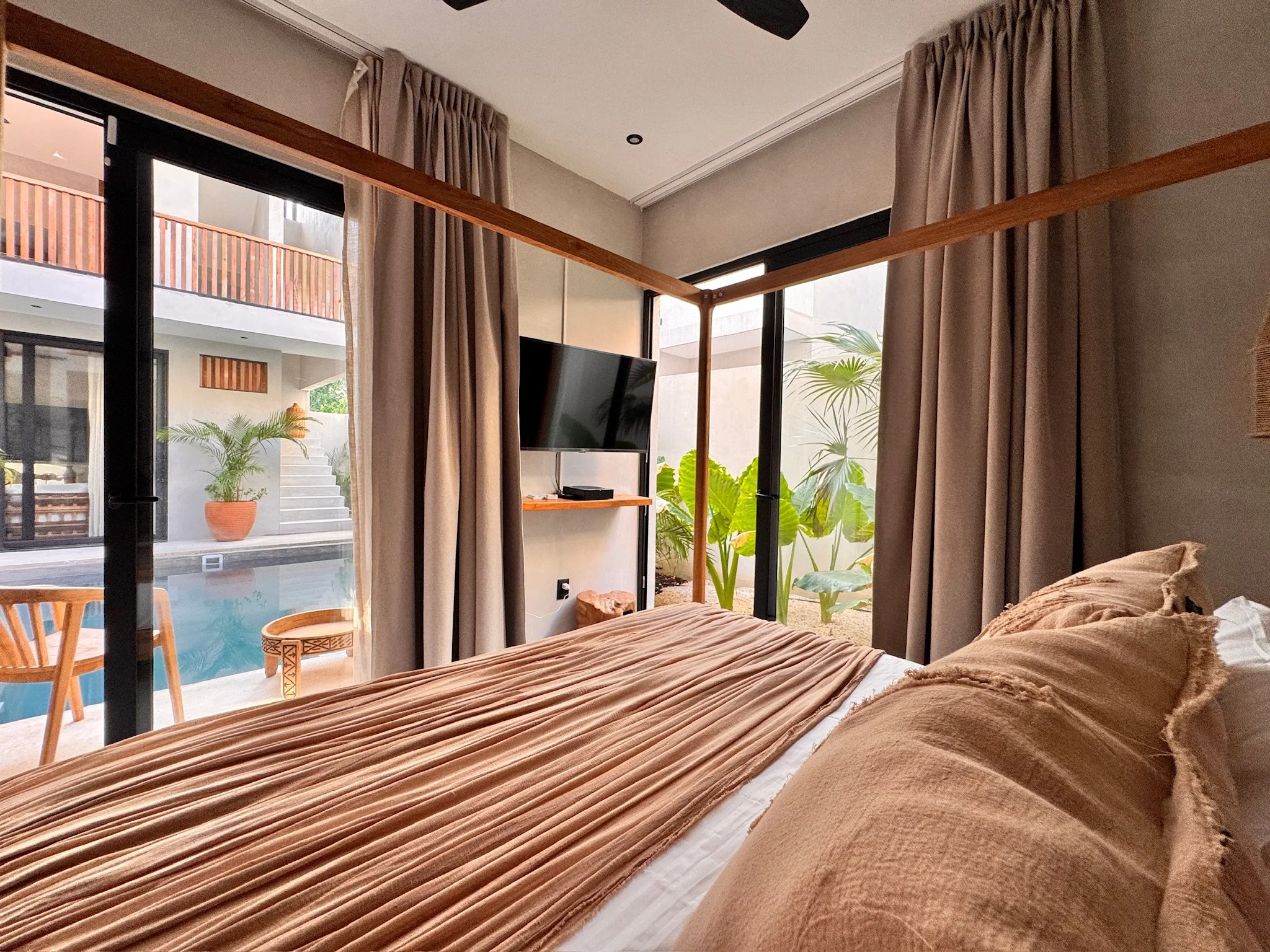 View of a bedroom with a bed, curtains, a wall-mounted TV, and large glass doors opening to an outdoor area with plants and a pool.