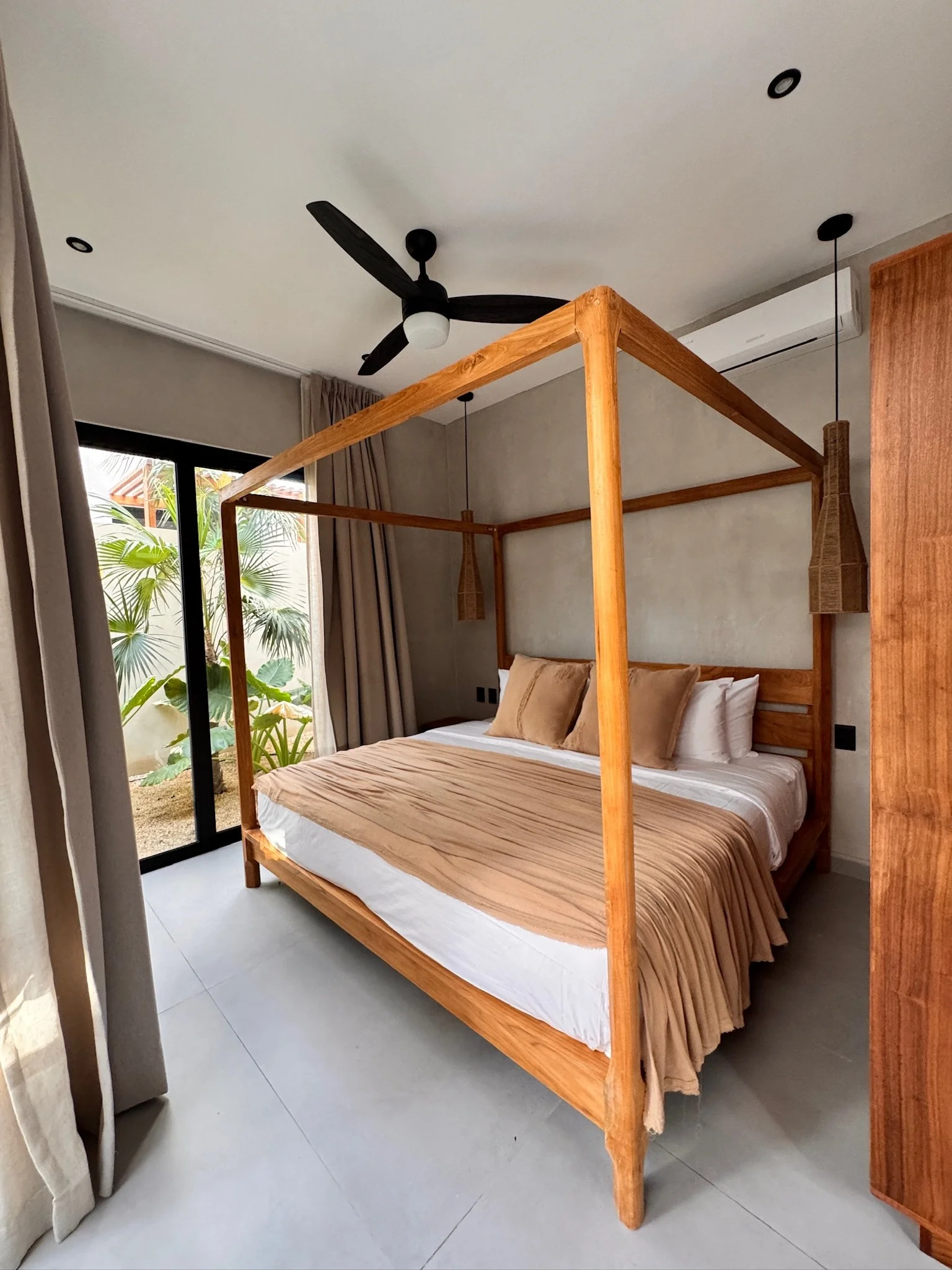 A bedroom with a wooden canopy bed, beige bedding, and pillows, next to a sliding glass door with curtains and outside greenery visible.