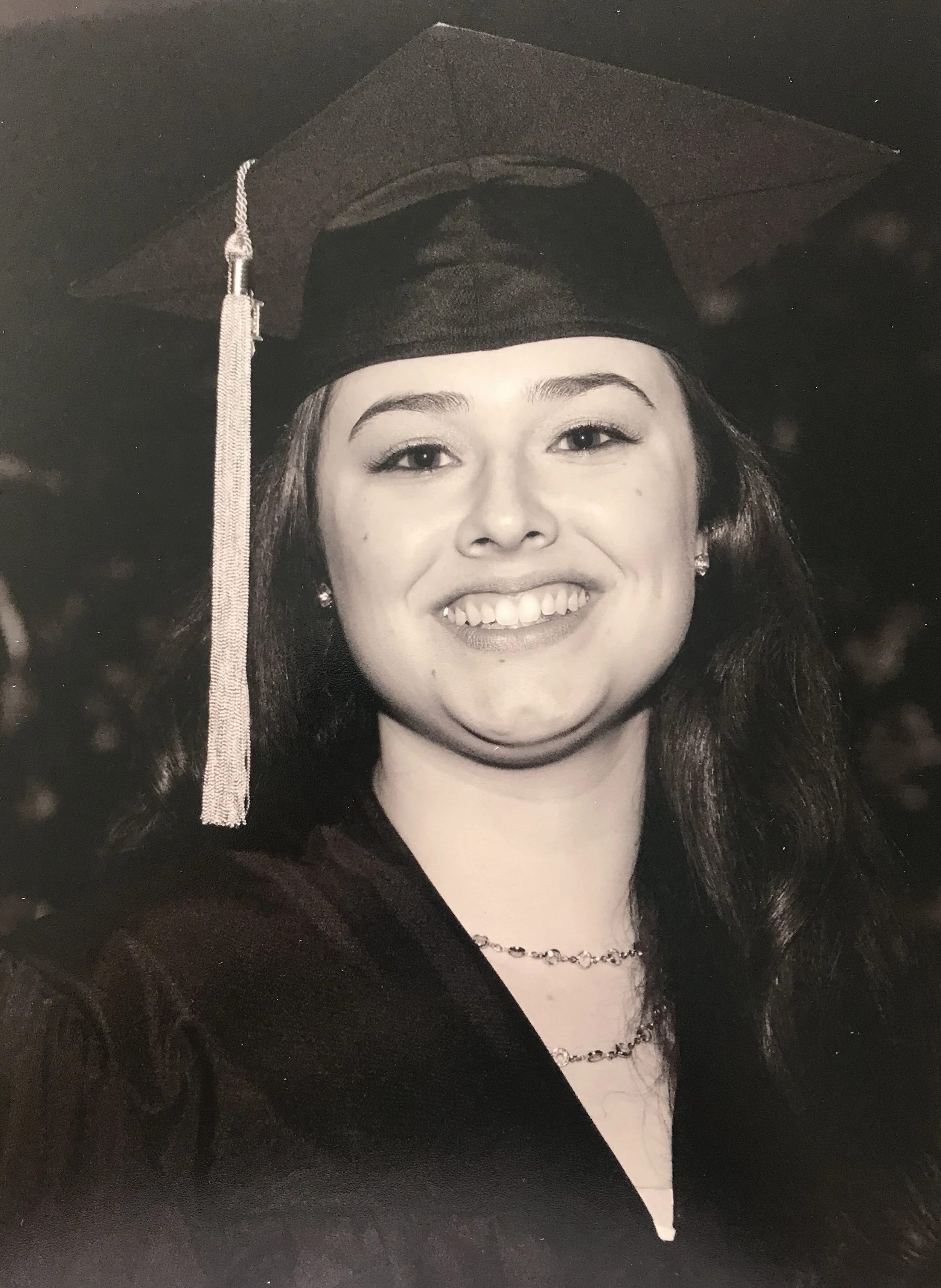 Black-and-white photo of a young woman in a graduation cap and gown, smiling, with a necklace visible on her neck.
