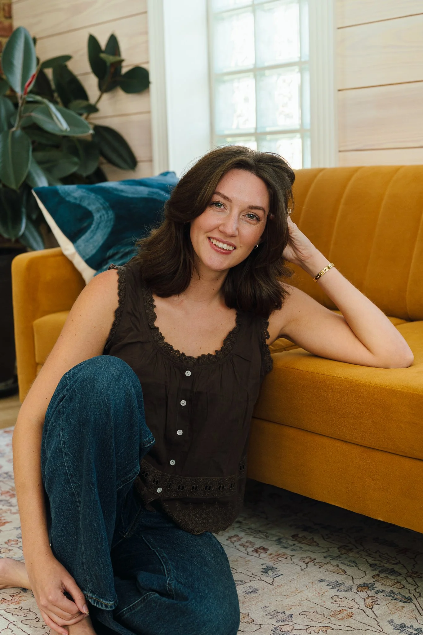 McLaine Beeman Mabry with shoulder-length brown hair, wearing a sleeveless dark top and blue jeans, is sitting on the floor beside a mustard yellow couch, smiling at the camera inside a cozy, wooden-paneled room.