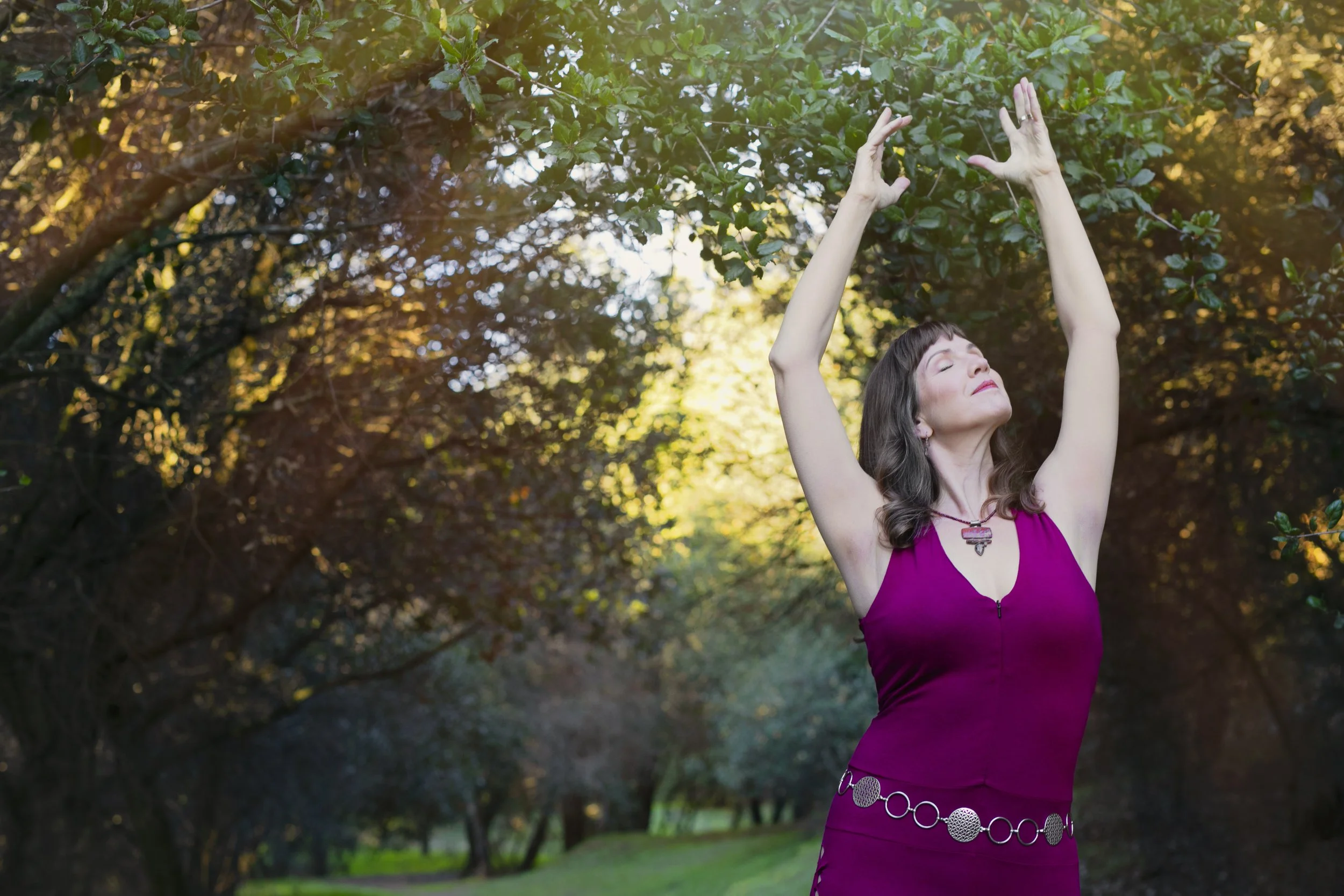 A woman with dark brown hair in a purple dress stands outdoors in a park, with her eyes closed and arms raised above her head in a relaxed pose, surrounded by trees and sunlight.