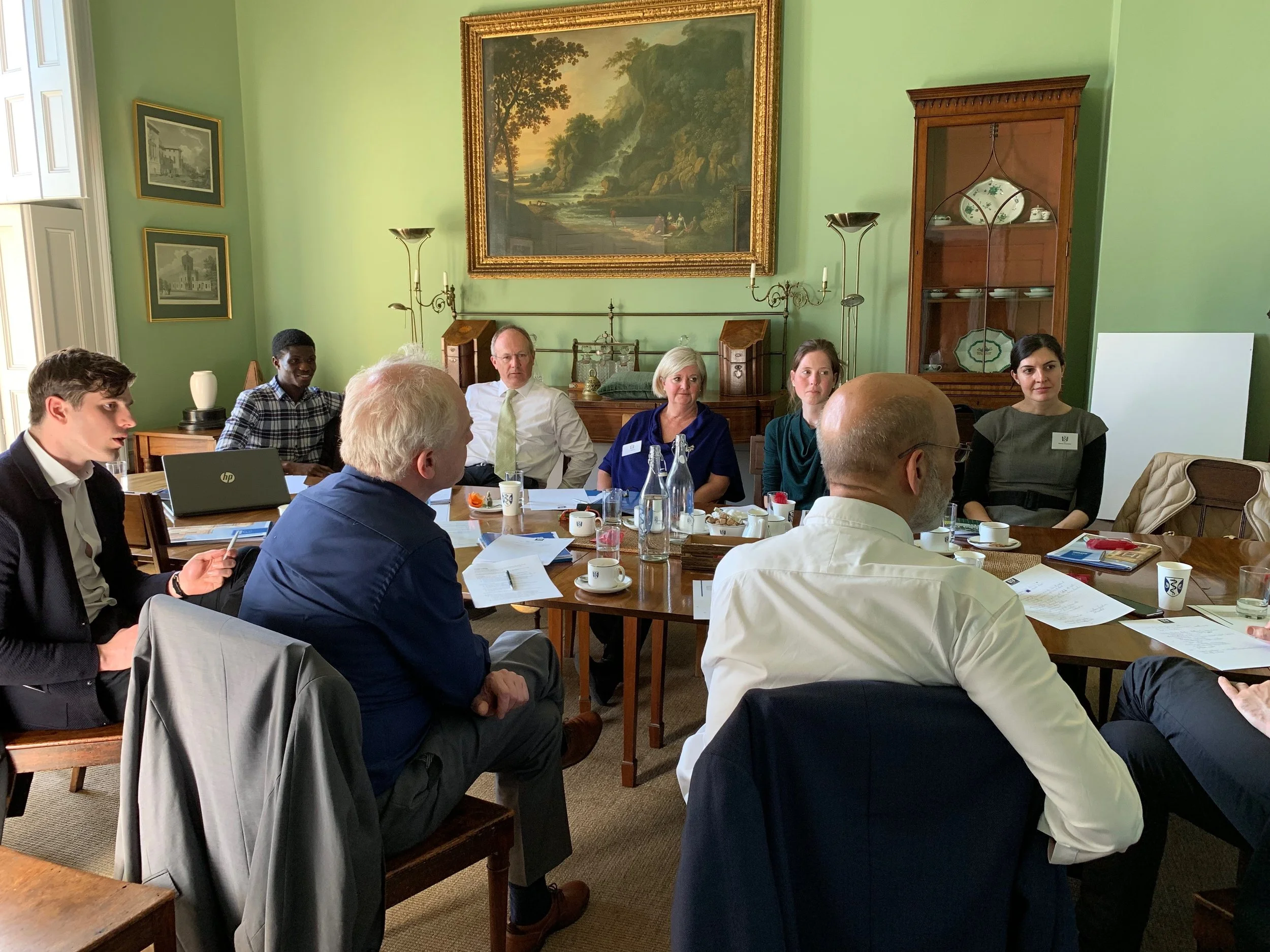 A group of diverse people sitting around a table in a formal meeting room in Oxford University. There are various paintings and a wooden cabinet on the walls. The table has coffee cups, water bottles, and papers.