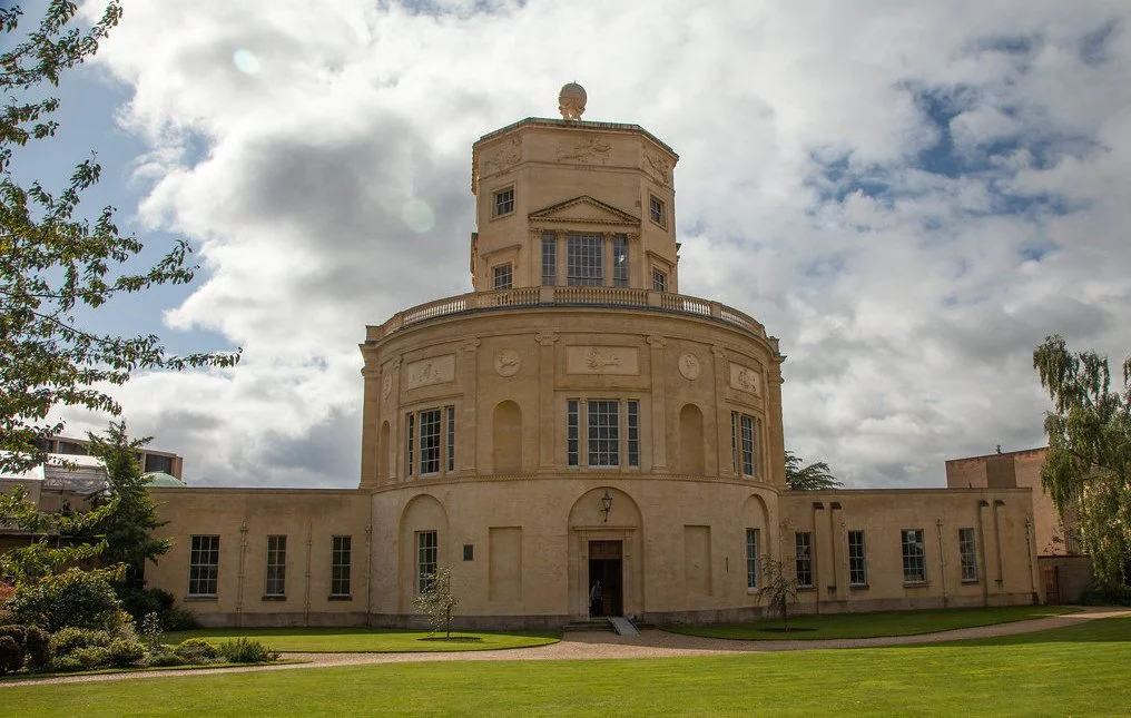 A beige, historic building with a rounded central tower, multiple windows, and a landscaped lawn in front under a partly cloudy sky. Green Templeton College, Oxford University.