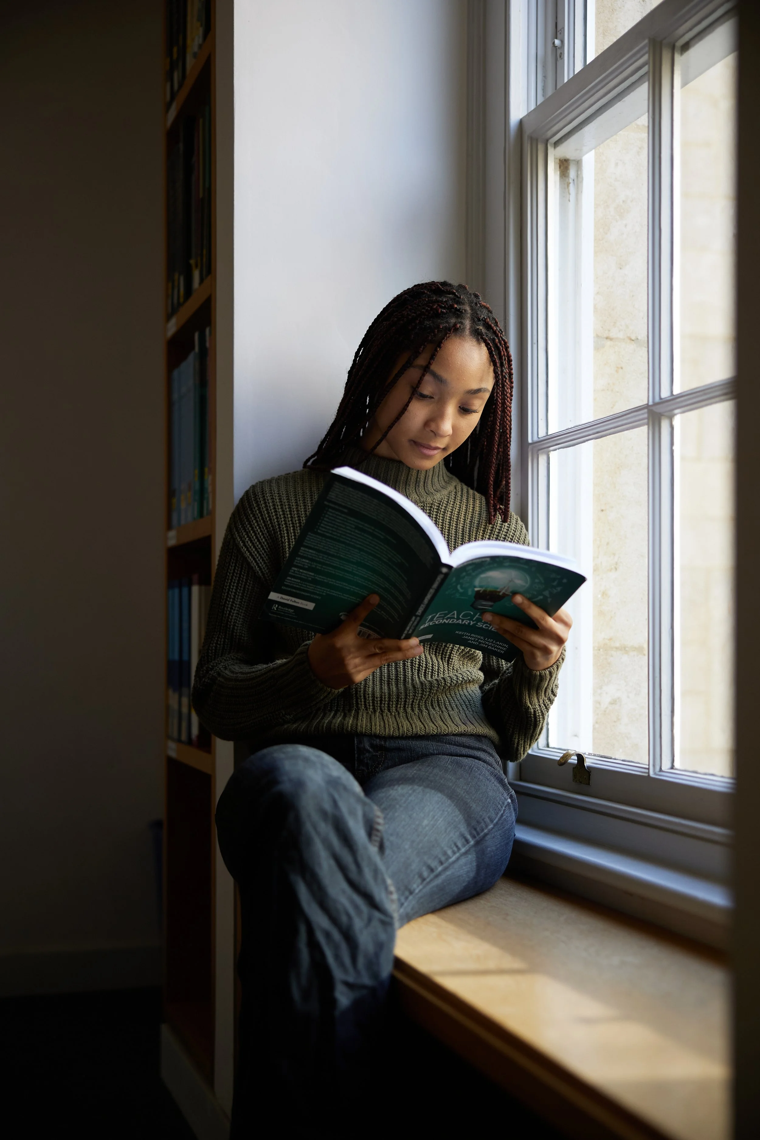 A young woman with braided hair is sitting by a window, reading a book, with her legs crossed on a windowsill. She is wearing a green striped sweater and jeans, and appears focused on her reading. At Oxford University.
