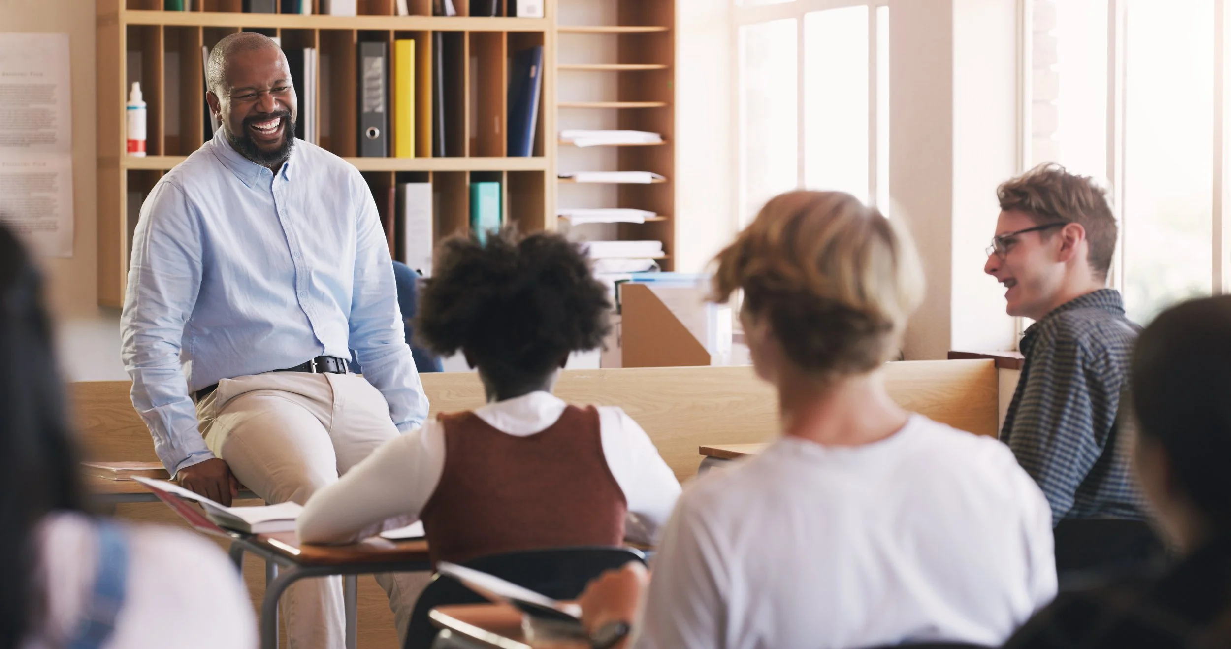 A diverse group of students sitting at desks in a classroom, smiling and listening to a male teacher who is leaning against the desk and laughing.