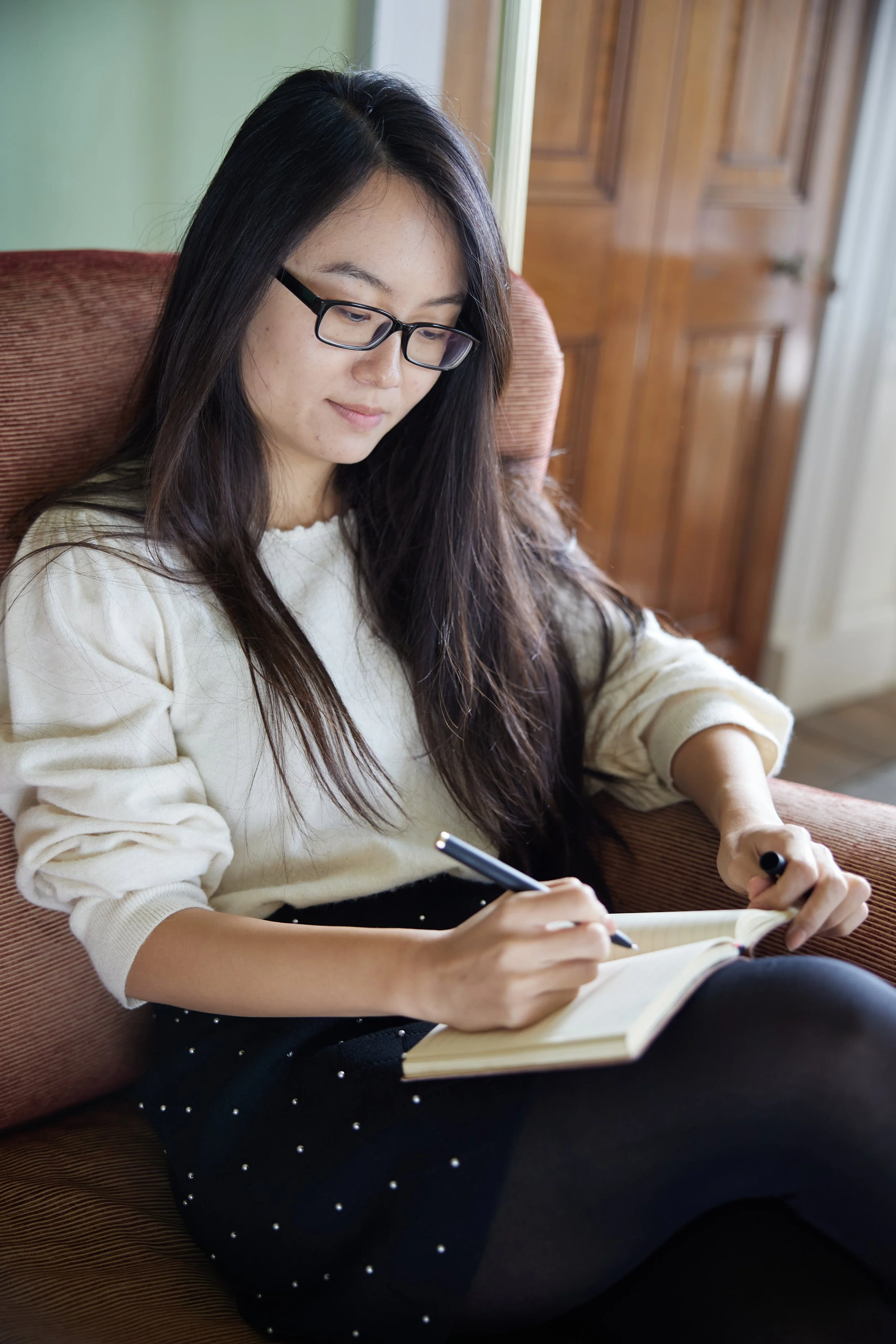 A young woman with long dark hair, glasses, wearing a light-colored sweater, sitting on a brown armchair, writing in a notebook with a pencil. At Oxford University
