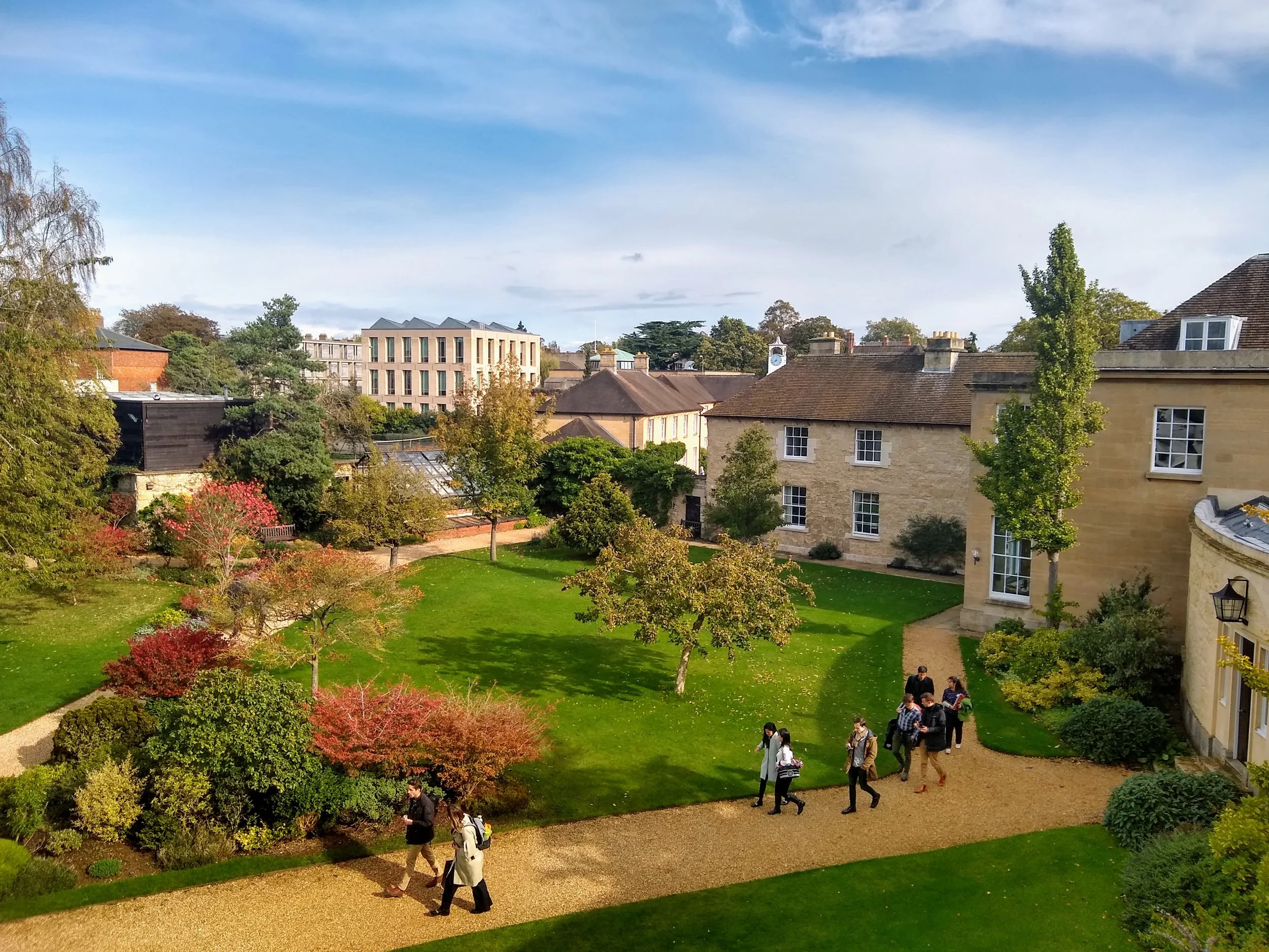 A garden courtyard with green grass, trees, and colorful plants, surrounded by historic and modern buildings, with people walking along gravel paths.
