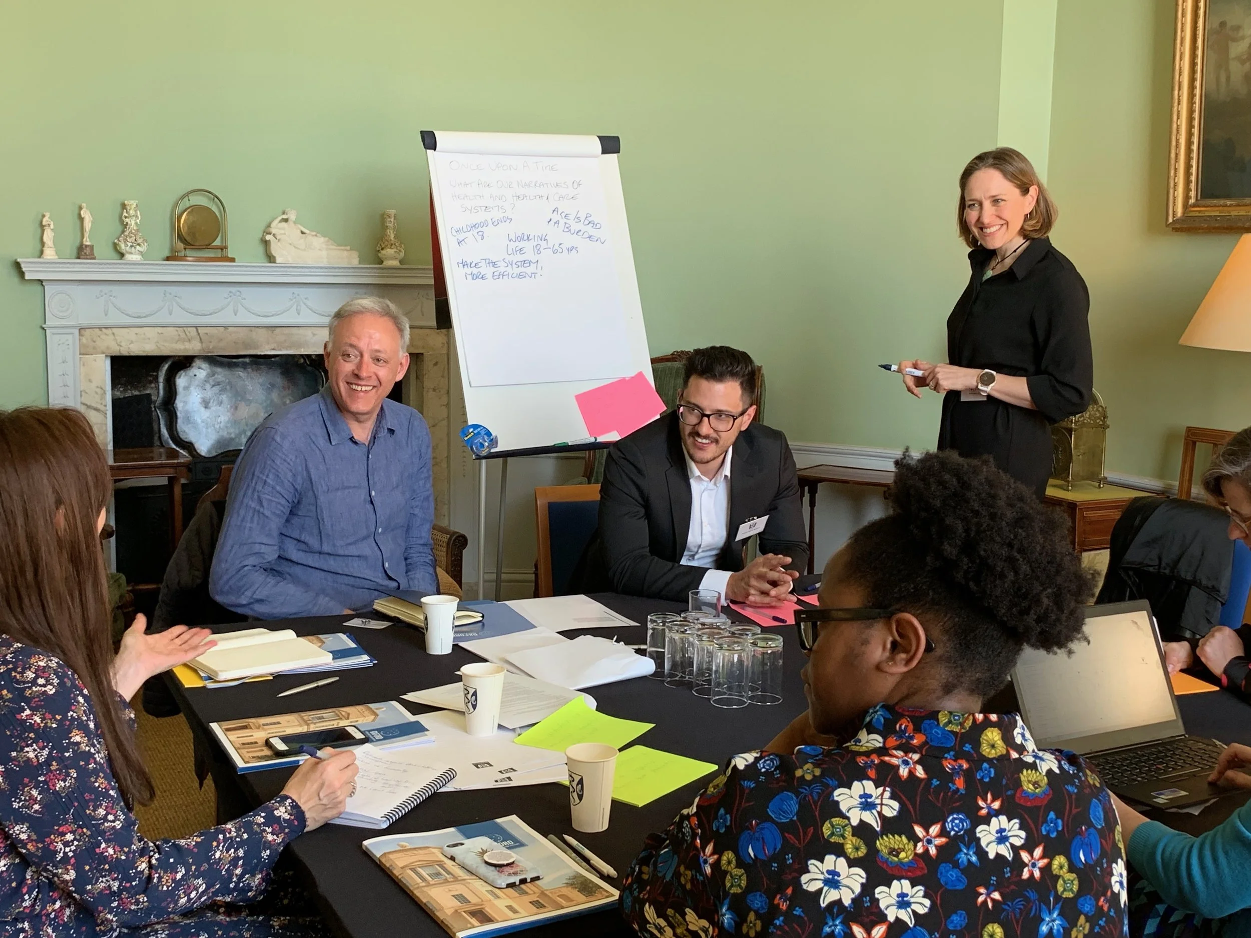 A group of people in a meeting or workshop. There is a woman standing and smiling, holding a marker near a flip chart. The flip chart has handwritten notes. Others are seated at a table with papers, notebooks, and cups.