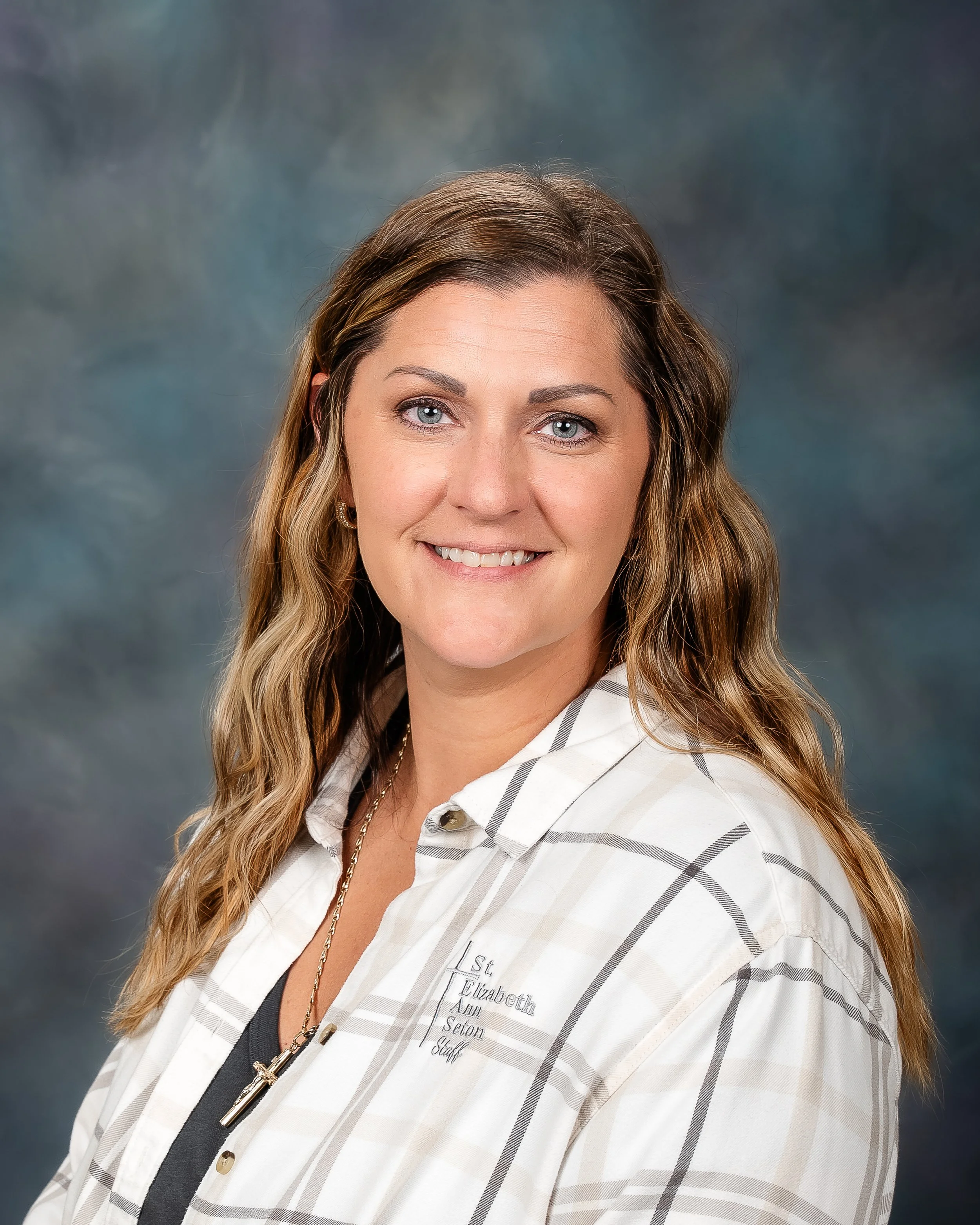 Headshot of a woman with long wavy brown hair, light blue eyes, wearing a white plaid shirt and a necklace with a cross pendant, against a dark blue-gray background.