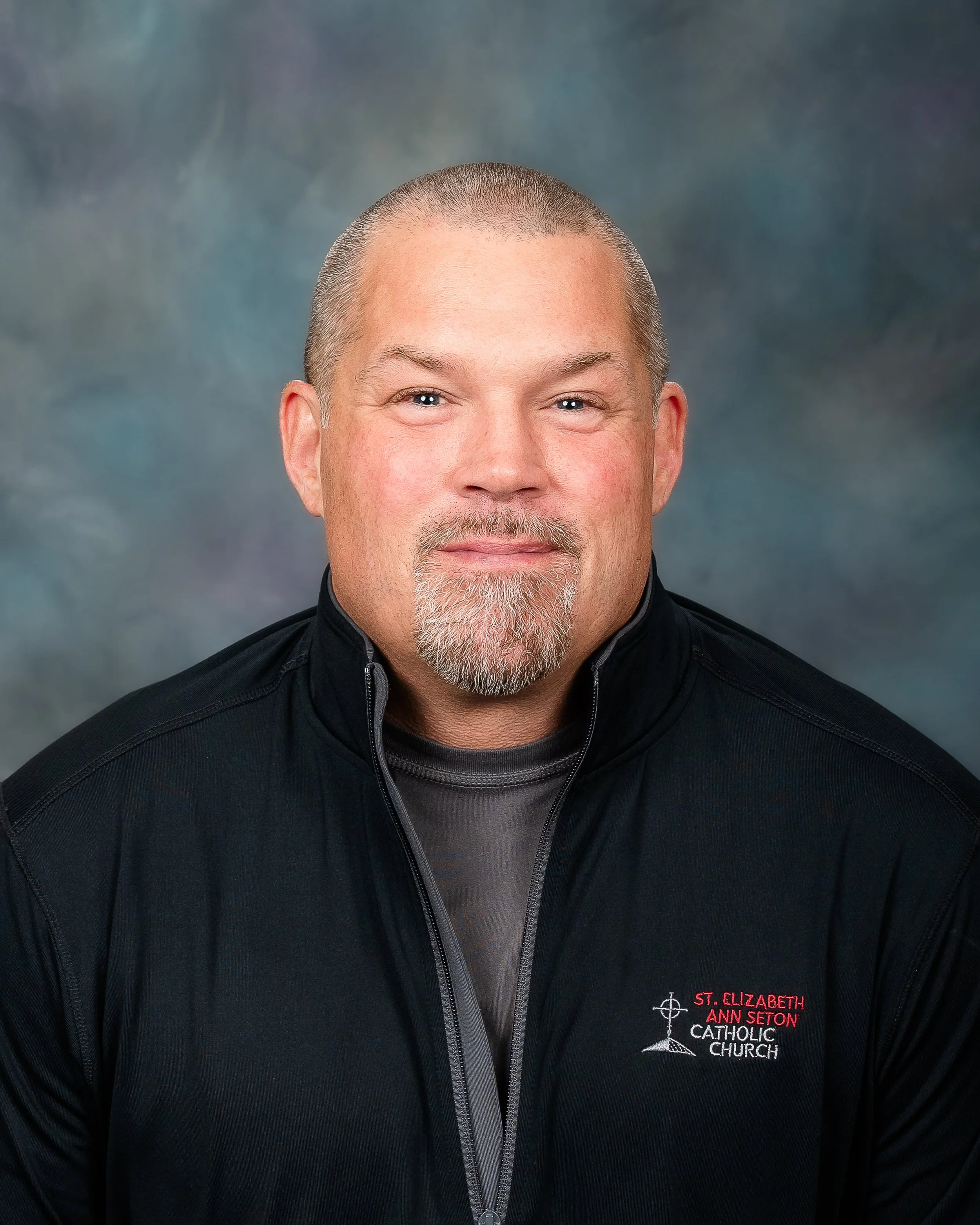 Man with short hair and a goatee wearing a black jacket with embroidery, posing for a professional portrait against a blurred background.