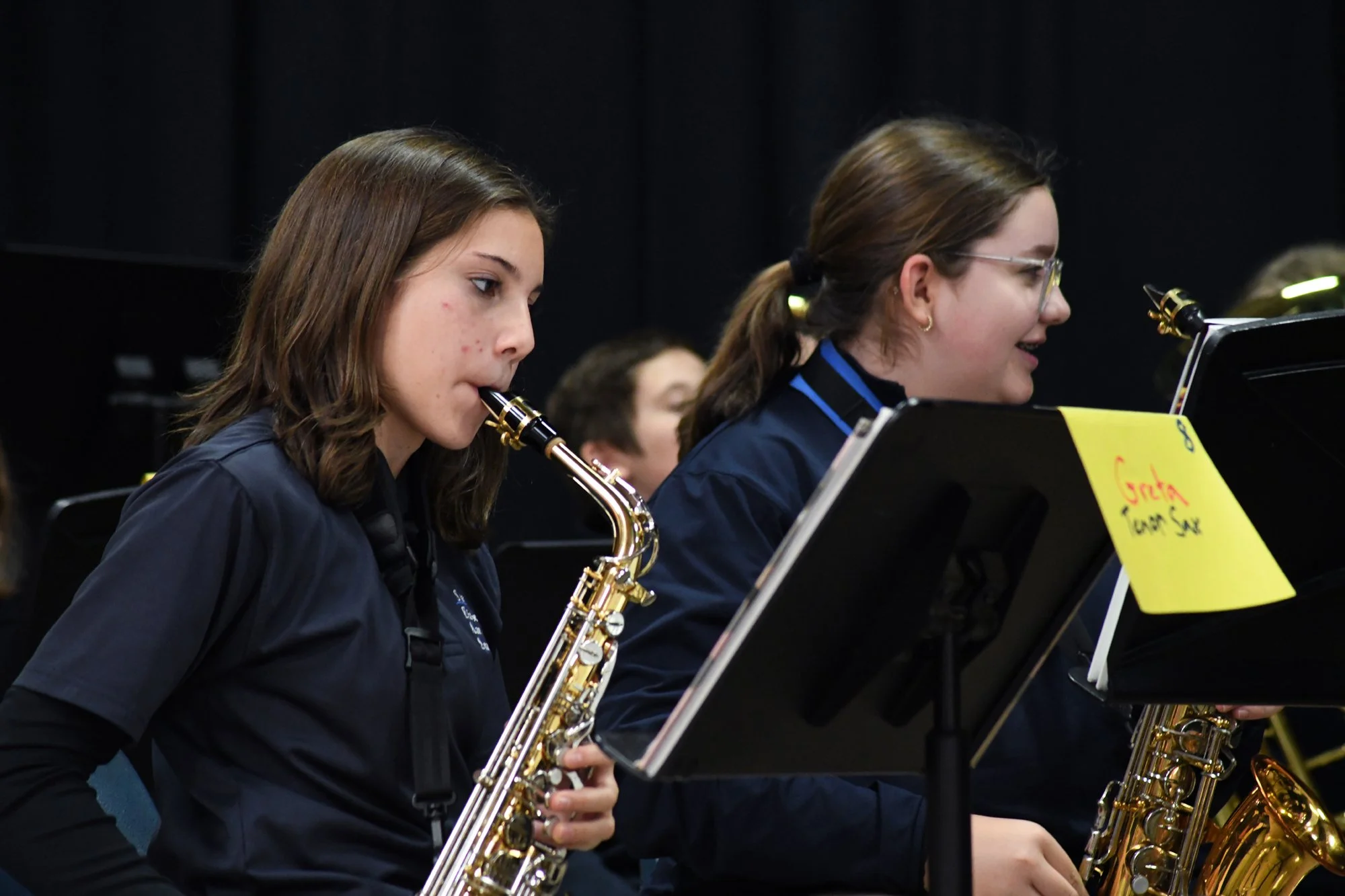 Two young women playing saxophones in a band, reading sheet music on music stands, in a performance setting.
