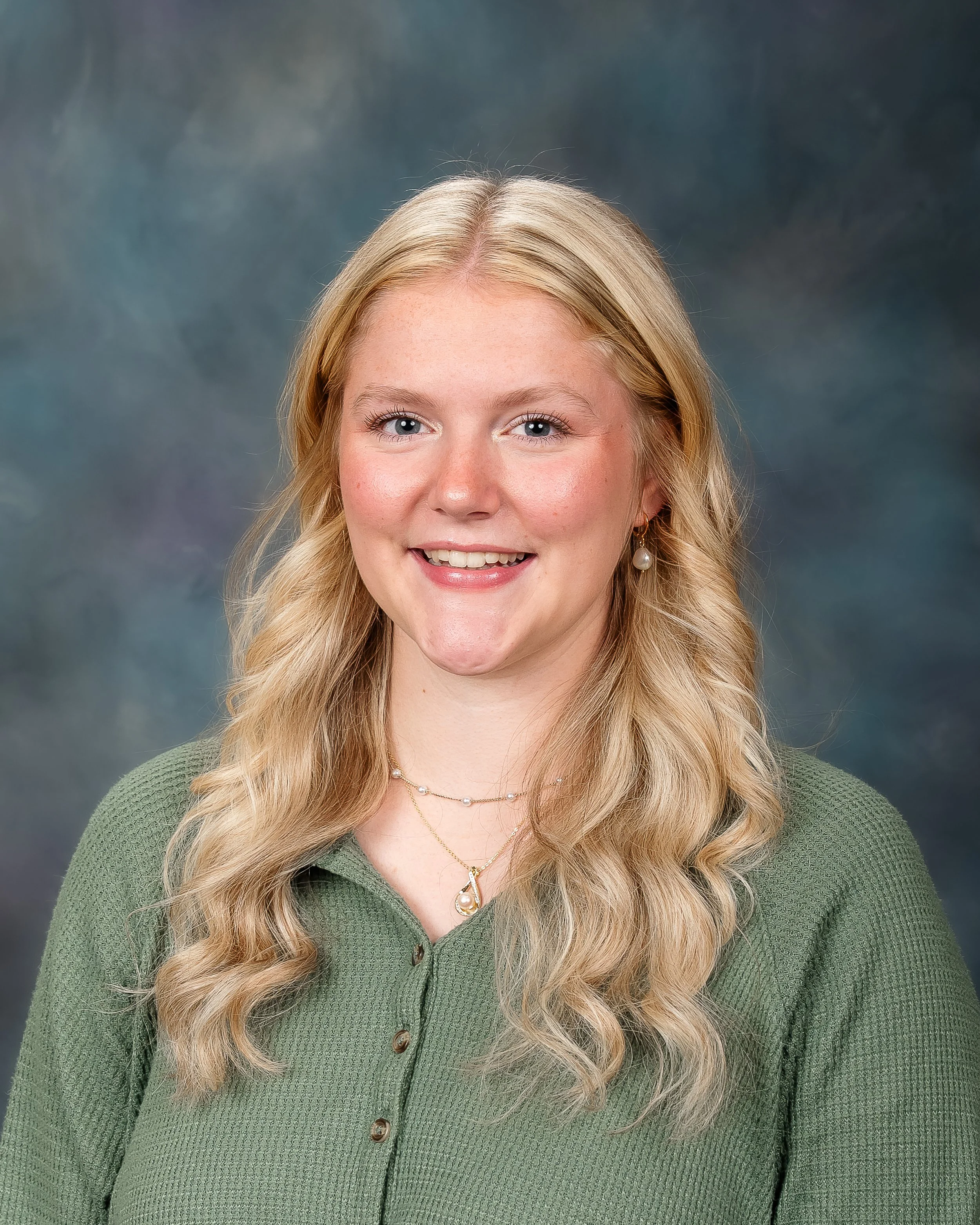 A smiling young woman with long wavy blonde hair, wearing a green button-up top and pearl earrings, standing against a colorful cloudy backdrop.