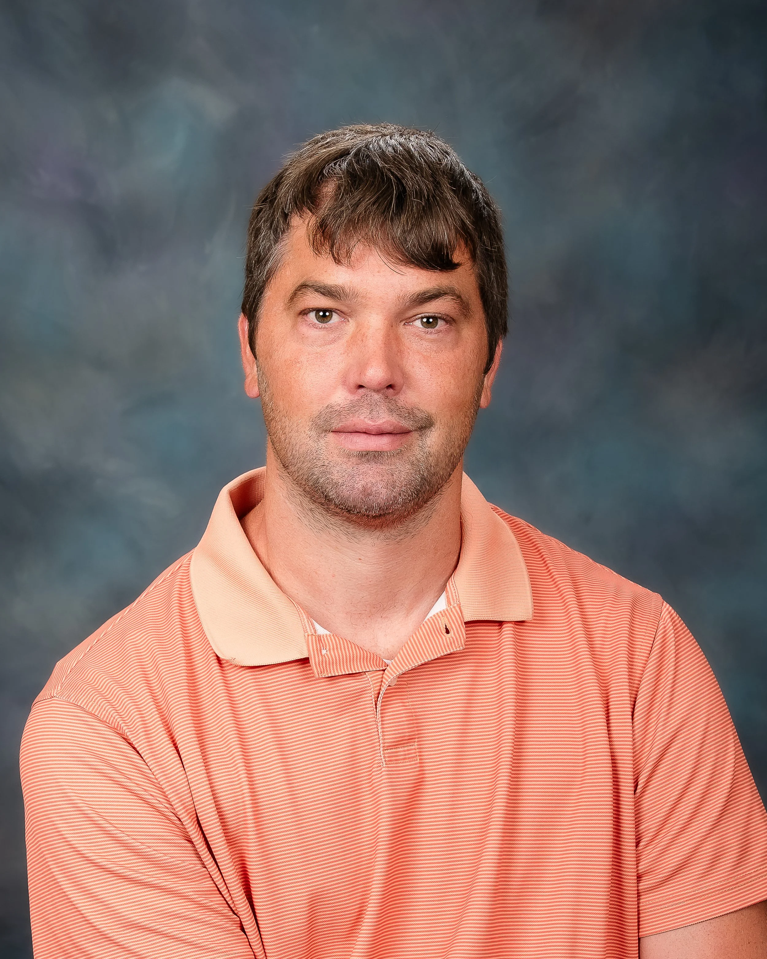 A portrait of a man with short brown hair, wearing a peach-colored, striped polo shirt, standing against a dark, cloudy background.