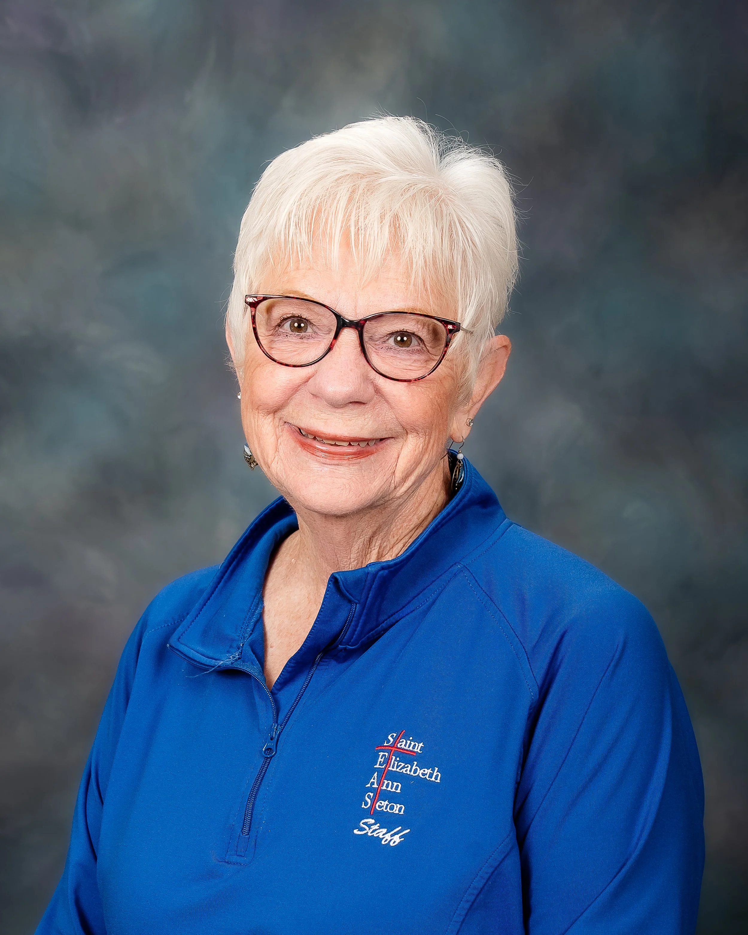 Senior woman with short white hair and glasses, wearing a blue zip-up jacket with embroidered text, smiling at the camera against a neutral background.
