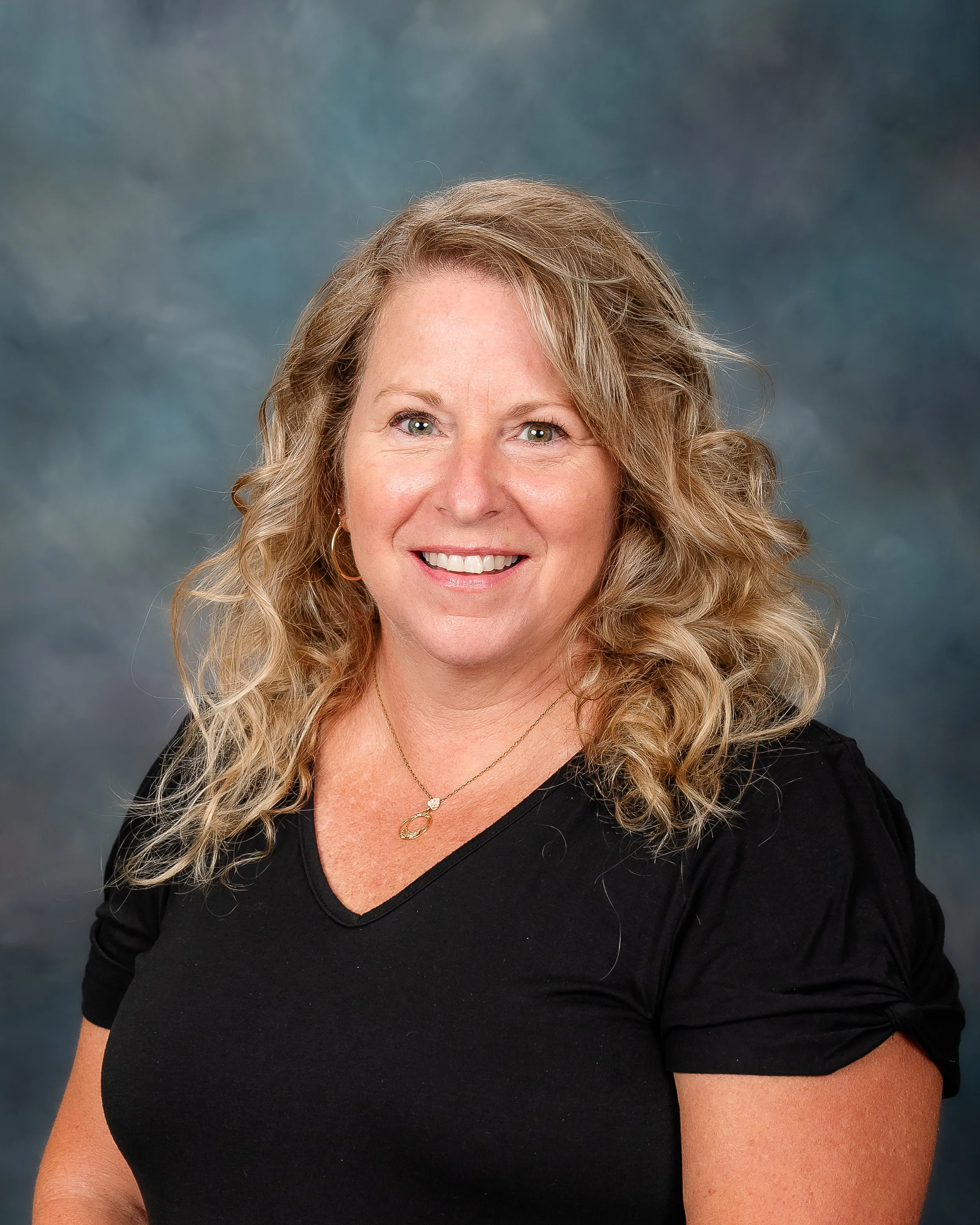 A woman with blonde curly hair smiling in front of a blue and gray blurred background.