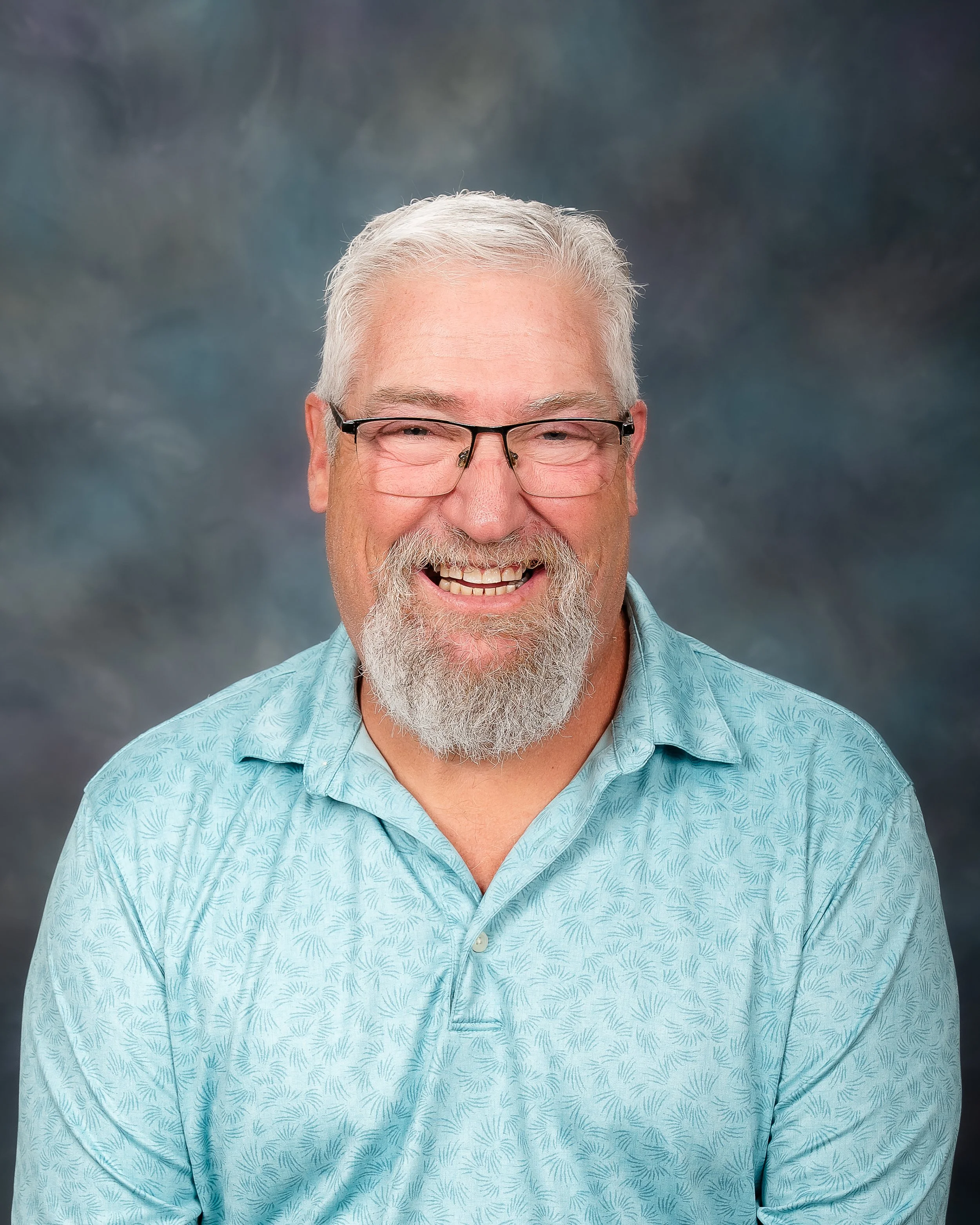 A smiling middle-aged man with gray hair, glasses, and a beard, wearing a light blue patterned shirt, against a blurred dark background.