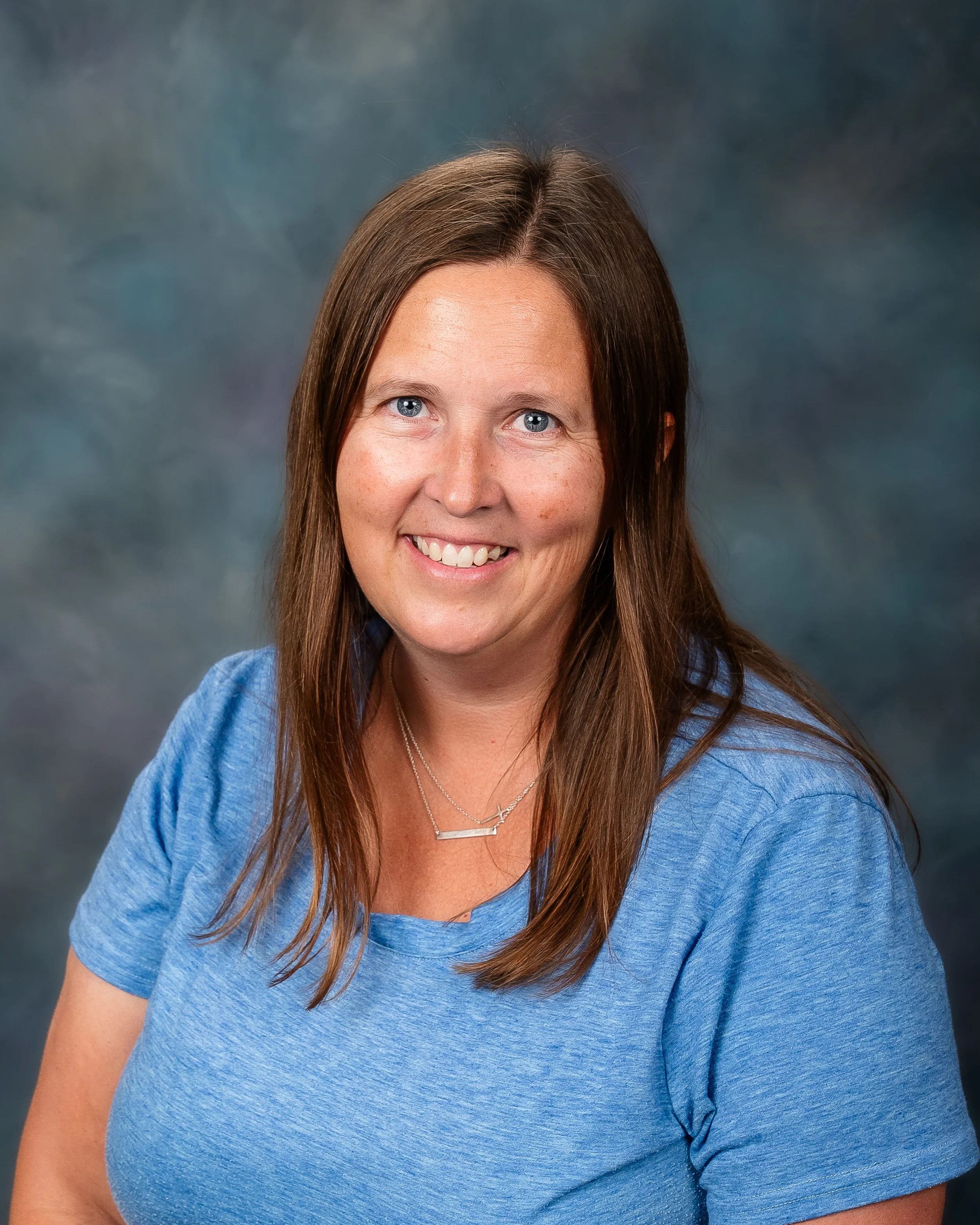 A woman with long brown hair, blue eyes, and a friendly smile, wearing a blue t-shirt and a silver necklace, poses against a greyish-blue background.
