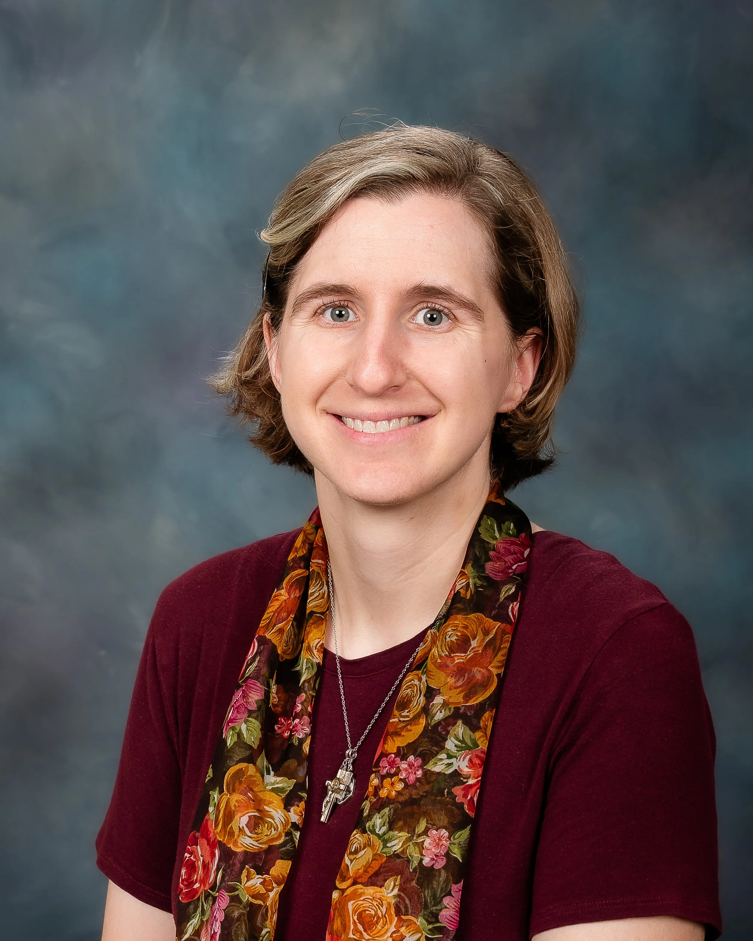 A woman with short, light brown hair and blue eyes, smiling and wearing a maroon shirt with a floral scarf and a necklace with a key pendant, posed against a multicolored background.