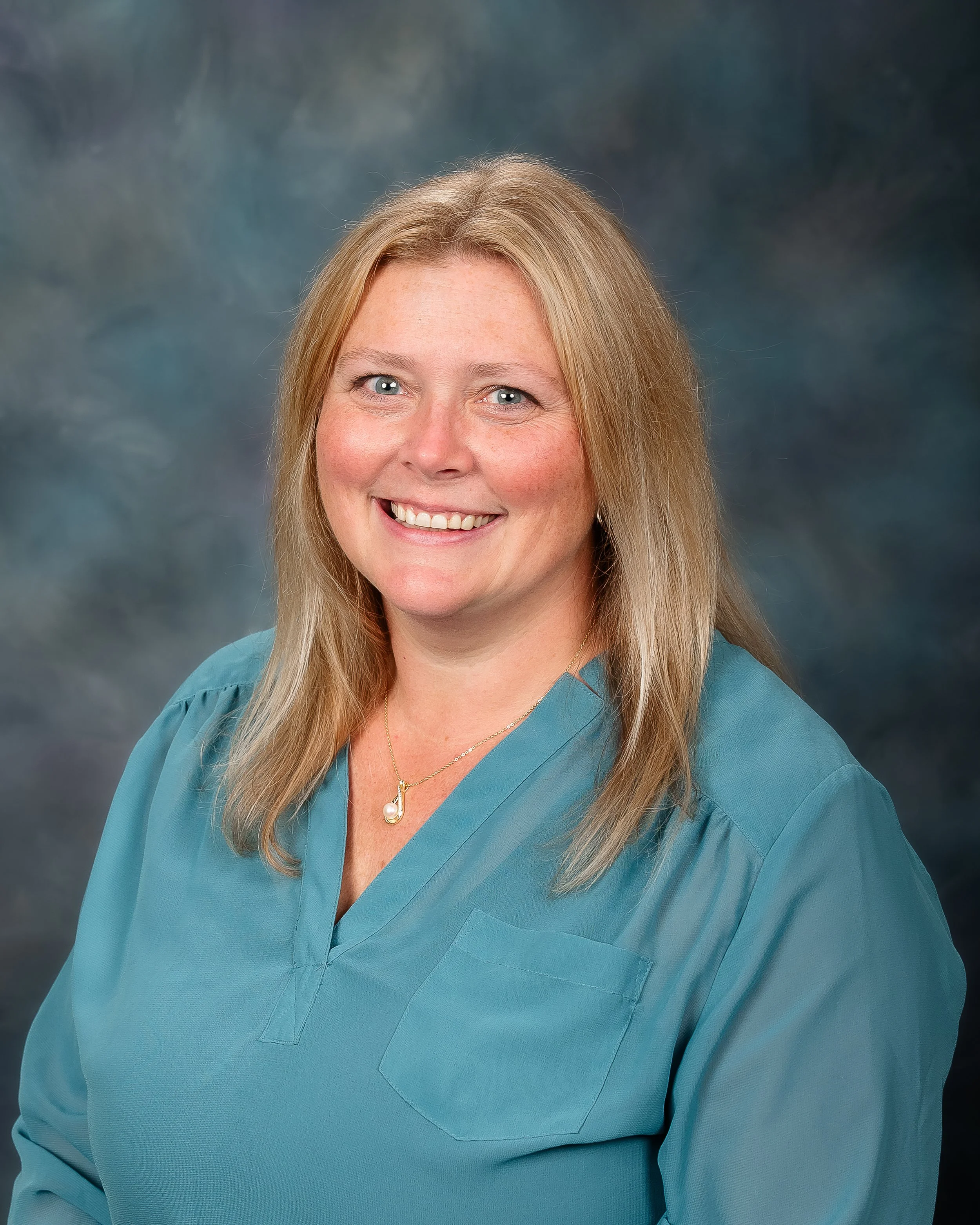 A woman with blonde hair smiling, wearing a teal medical uniform and a gold necklace, against a dark, cloudy background.