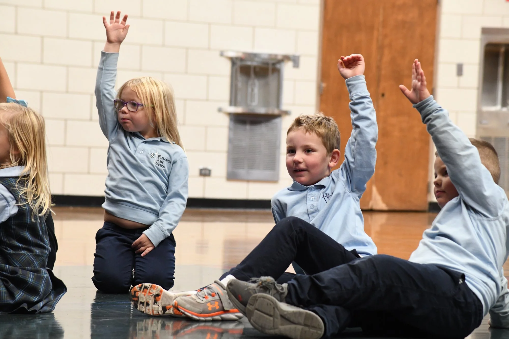 Three young children in school uniforms with light blue shirts sitting on the floor in a classroom, raising their hands for a question or to participate in an activity.