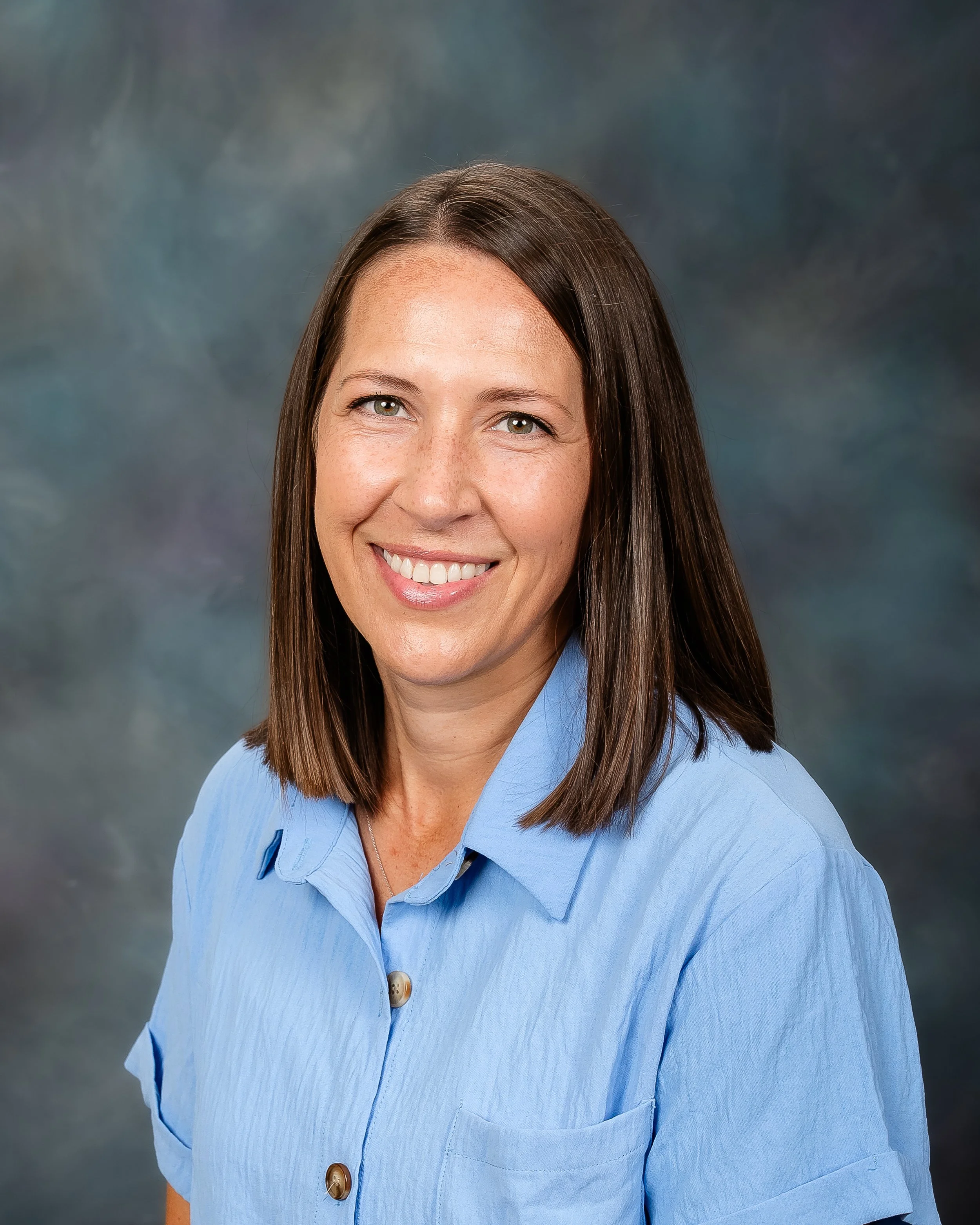 A woman with medium-length brown hair smiling and wearing a light blue collared shirt against a blurred, multicolored background.