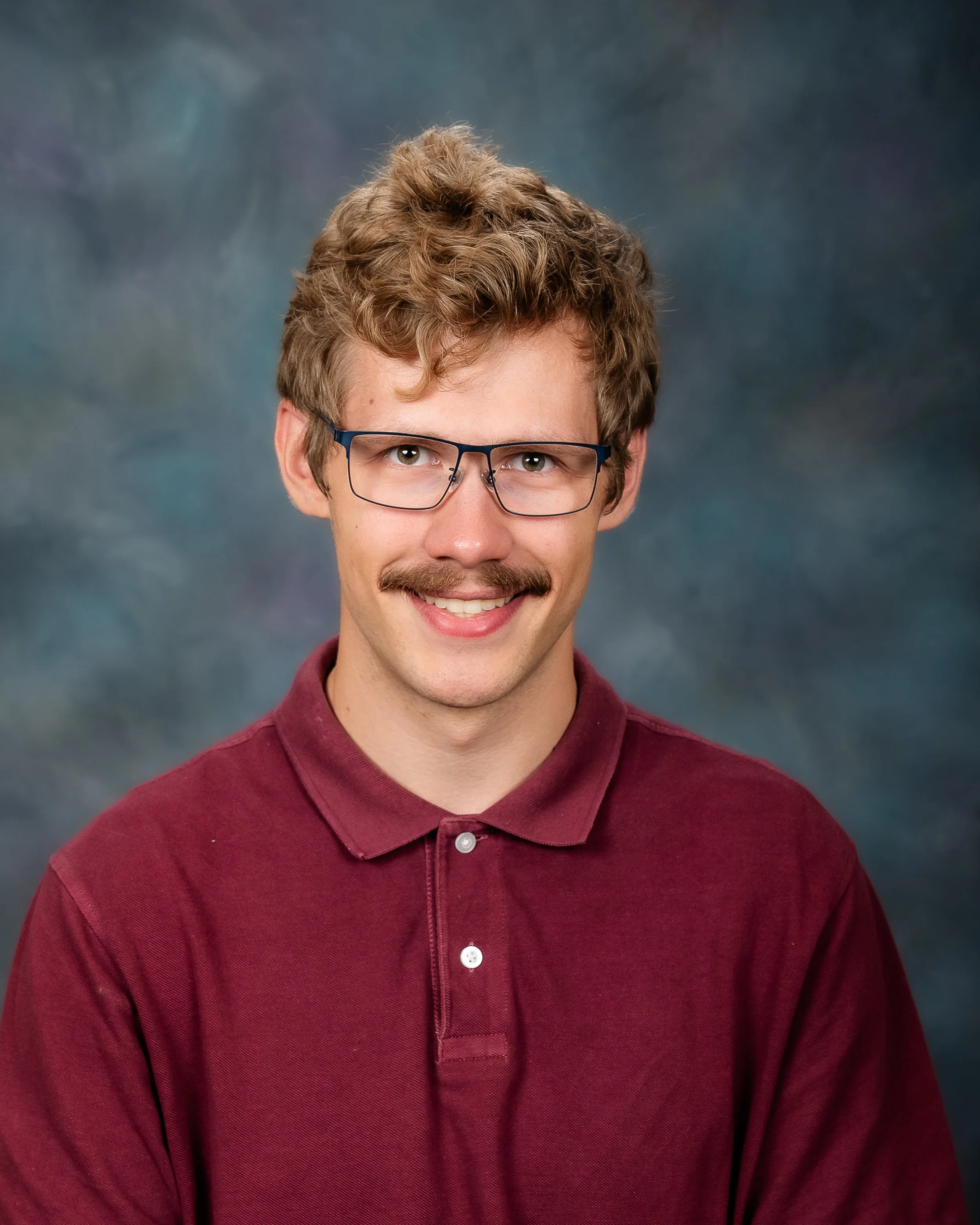 A man with light brown curly hair, glasses, a mustache, and wearing a maroon polo shirt, smiling in front of a dark, cloudy background.