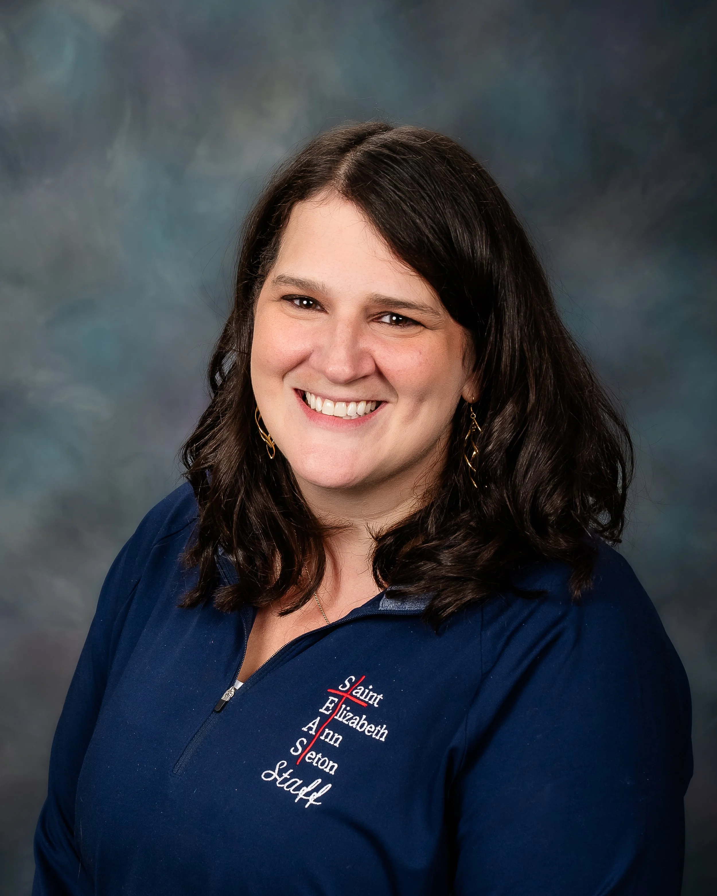 A woman with dark brown hair smiling, wearing a navy blue jacket with 'Saint Elizabeth Ann Seton Staff' embroidered on it, against a blurred colorful background.