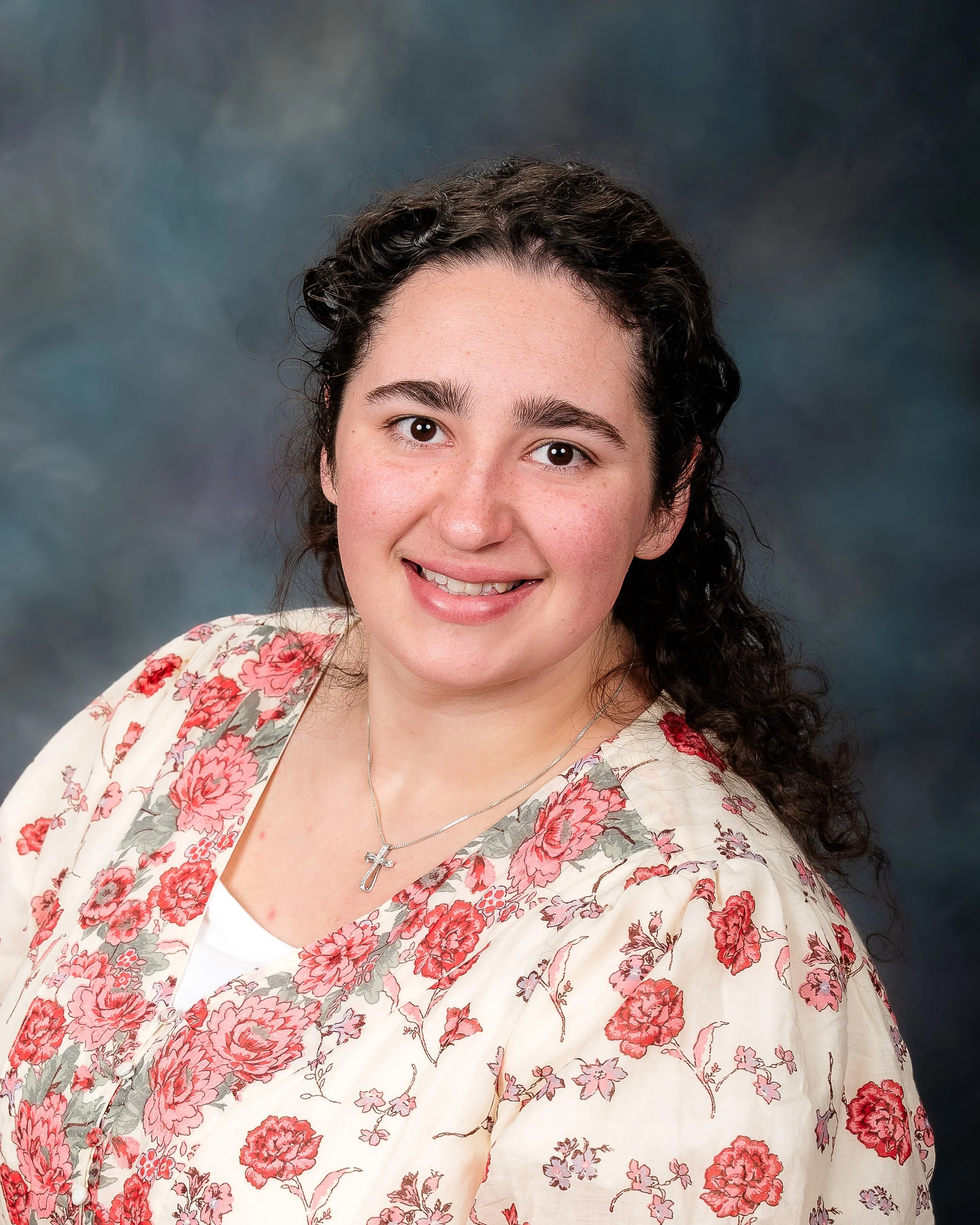 A woman with curly dark hair and a friendly smile, wearing a floral blouse and a silver cross necklace, against a dark, cloudy background.
